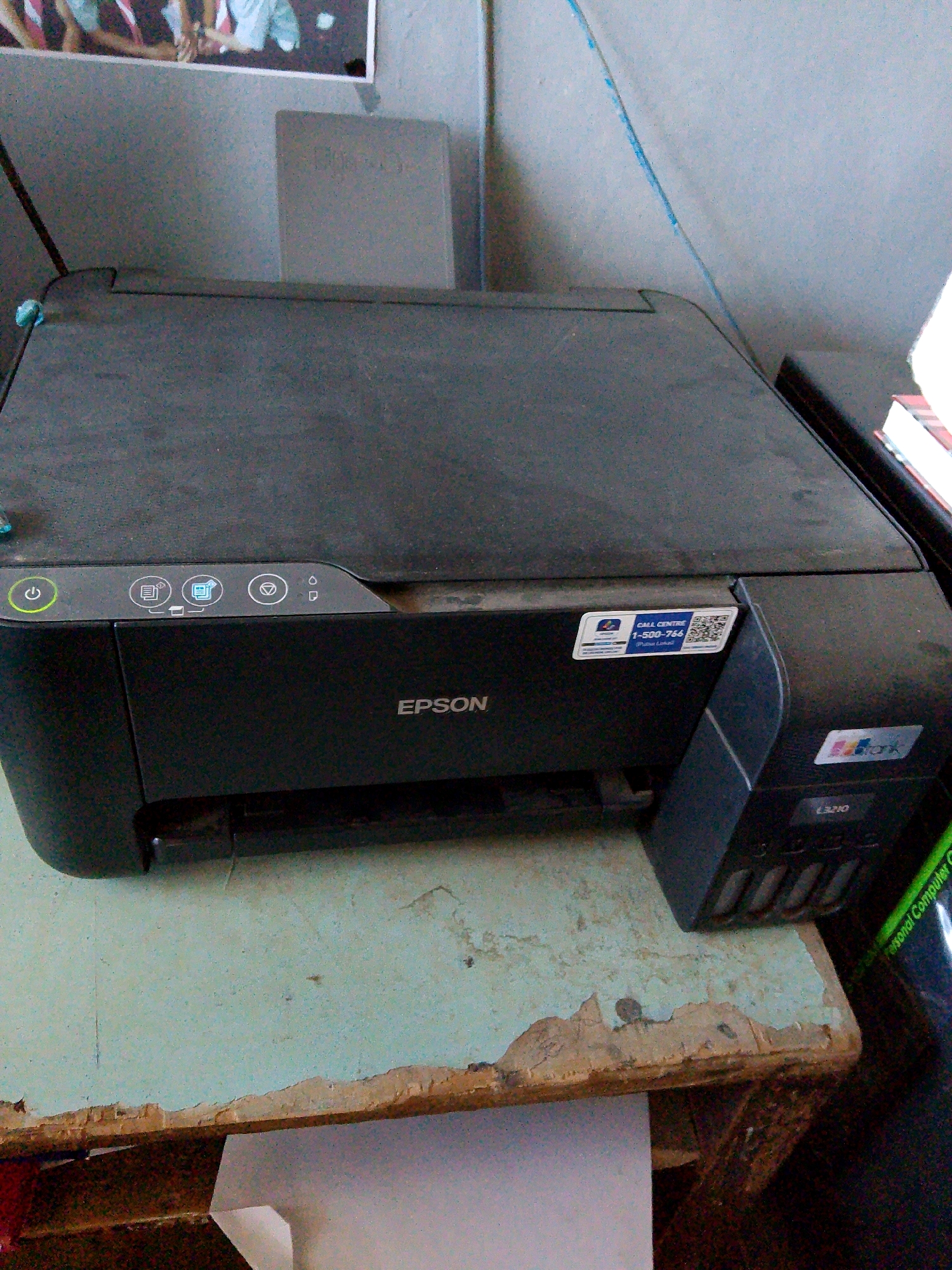 The image shows an Epson printer on a worn, light blue desk. The printer is black and has a flatbed scanner on top. There are several buttons on the front panel, including a power button, buttons for scanning, copying, and other functions. A sticker with "CALL CENTRE 1-800-766" and a QR code is affixed to the right side of the printer. Below the printer, there are visible ink tanks. The desk is made of wood and shows signs of age and wear, with chipped paint and exposed wood grain. In the background, a framed picture of people is visible on a gray wall. The overall setting appears to be an indoor workspace, possibly an office or home. The specific location in Kuripan Utara, Indonesia, cannot be determined from the visual information alone.