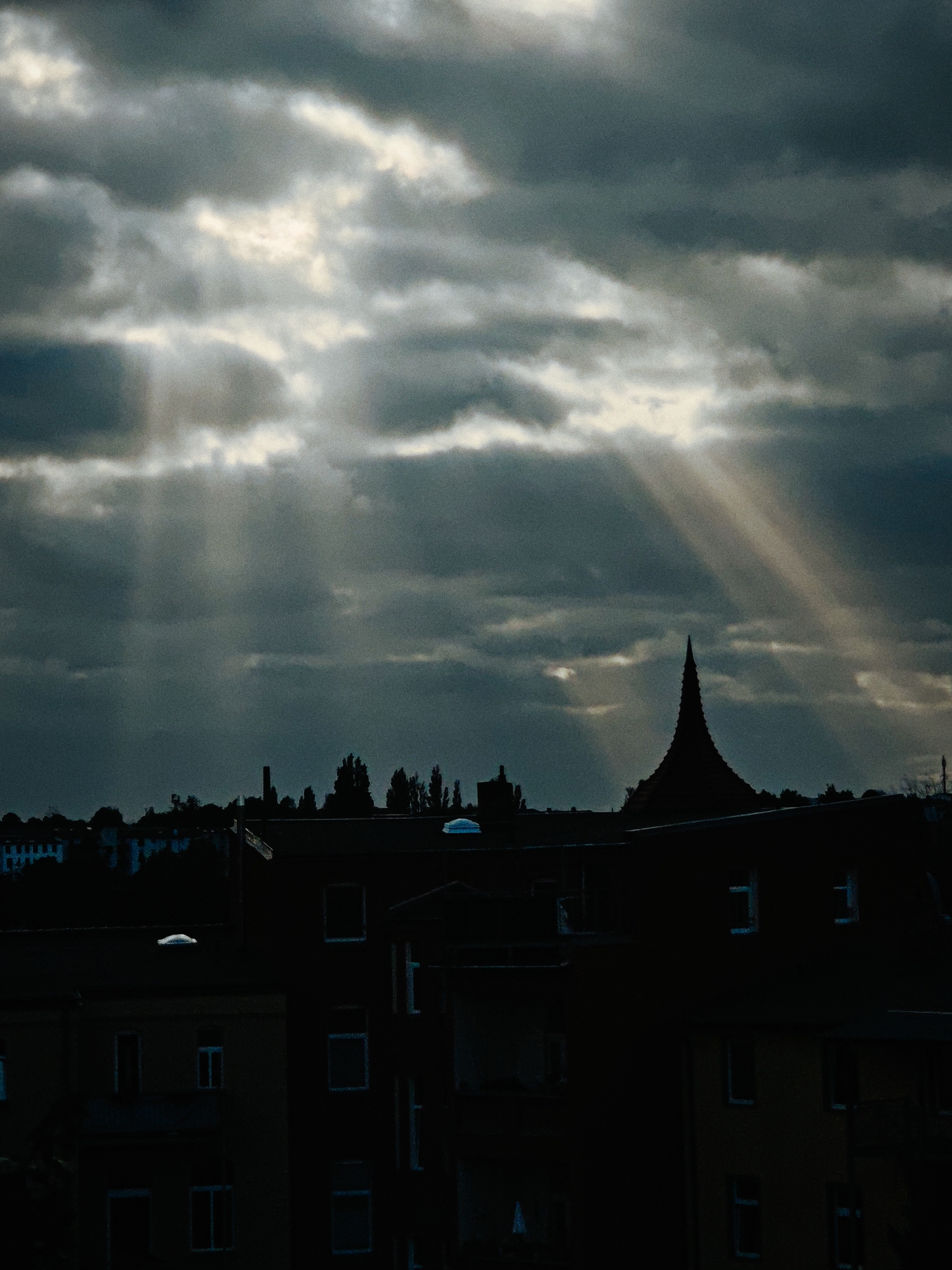 The image captures a dramatic sky over the city of Schwerin, Germany. Thick, dark clouds dominate the upper half of the frame, with shafts of sunlight piercing through them, creating a crepuscular effect. These light rays are angled, suggesting a low sun angle, possibly during sunrise or sunset. Below the sky, a silhouetted cityscape is visible. The architecture appears to be predominantly residential buildings, characterized by dark facades and visible window frames. One distinctive structure with a pointed, conical roof stands out in the middle ground, silhouetted against the sky. Trees and other buildings form a dense line along the horizon, also rendered in dark silhouette. The overall mood is somber and atmospheric, accentuated by the contrast between the dark clouds and the bright sunbeams. The scene is devoid of any visible people or distinct actions, focusing entirely on the natural light and urban landscape.