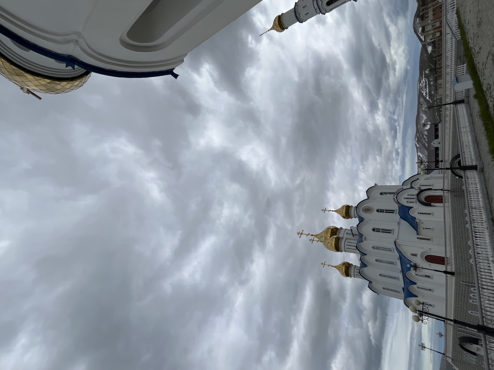 The image displays the Trinity Cathedral in Petropavlovsk-Kamchatsky, Russia, under a heavily overcast sky. The white, multi-domed cathedral, adorned with gold cupolas and blue trim, dominates the left side of the frame. In the foreground, a white balustrade and lampposts line a paved walkway. To the right, part of another white structure, possibly a bell tower, is visible, also topped with a gold dome. In the background, a town with various buildings and snow-capped mountains can be seen. No people are present in the image.