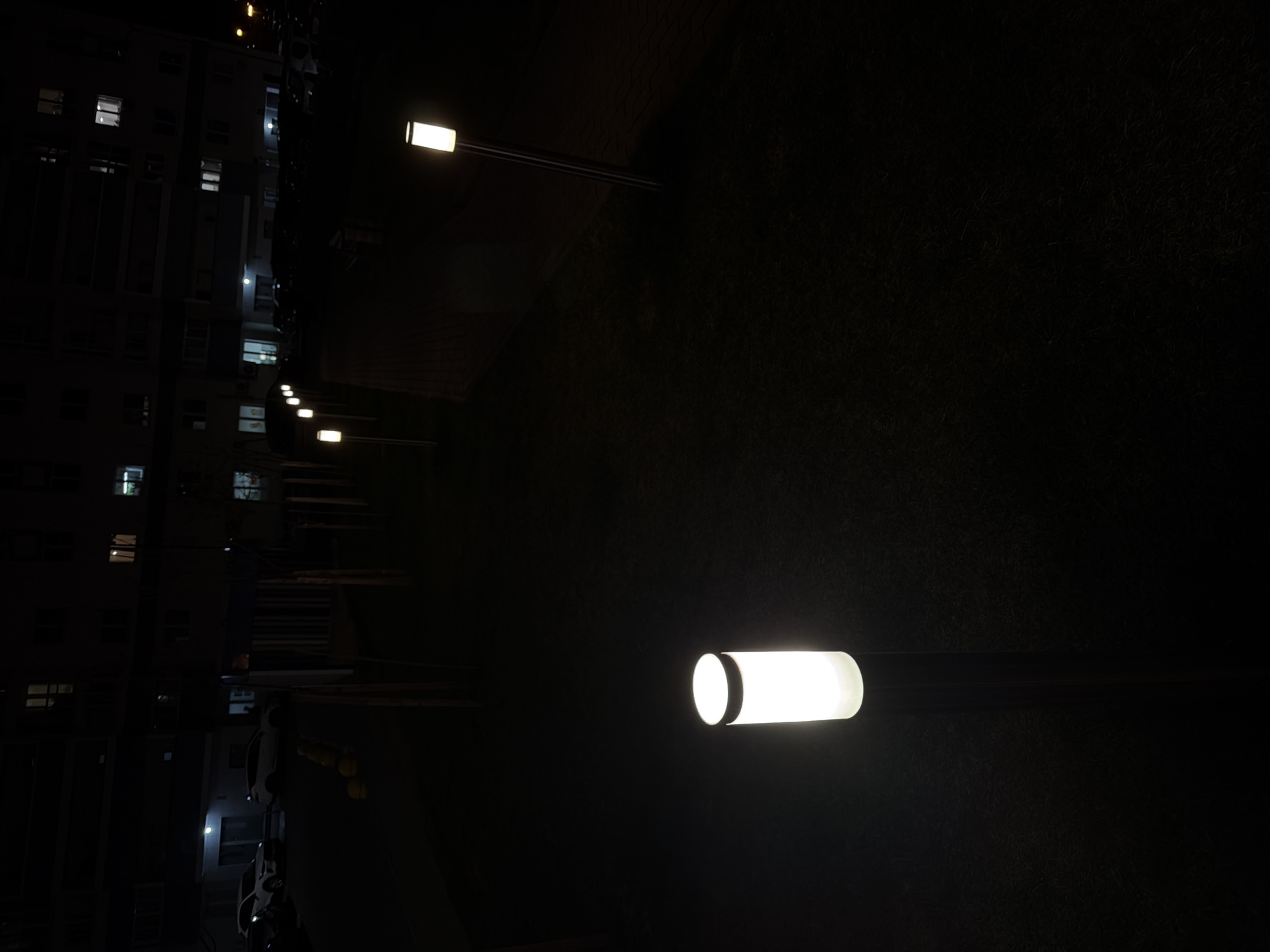 The media file captures a nocturnal outdoor scene in Minsk, Belarus, dominated by darkness punctuated by artificial light sources. The primary subjects are several modern streetlights with cylindrical fixtures, illuminating small portions of the environment. The closest and brightest streetlight is positioned in the lower right foreground, casting a strong white glow. A line of similar, though dimmer, streetlights recedes into the left background, creating a perspective of depth.
To the left of the image, a multi-story building is partially visible, appearing dark and silhouetted against the night sky. Many of its windows are illuminated, indicating internal activity or occupied units. Below the building, some indistinct shapes might be parked vehicles, but the darkness obscures clear identification. The ground occupying the right and central portions of the image is largely obscured by shadow, appearing as a dark, textured expanse, possibly grass or rough terrain.
The scene depicts a quiet, static moment at night. There are no people visible, and no discernible activity is taking place. The weather appears clear, with no visible precipitation or atmospheric haze. The overall mood is calm and somewhat stark, defined by the contrast between the bright artificial lights and the deep shadows of the surroundings. No visible text is present in the image.