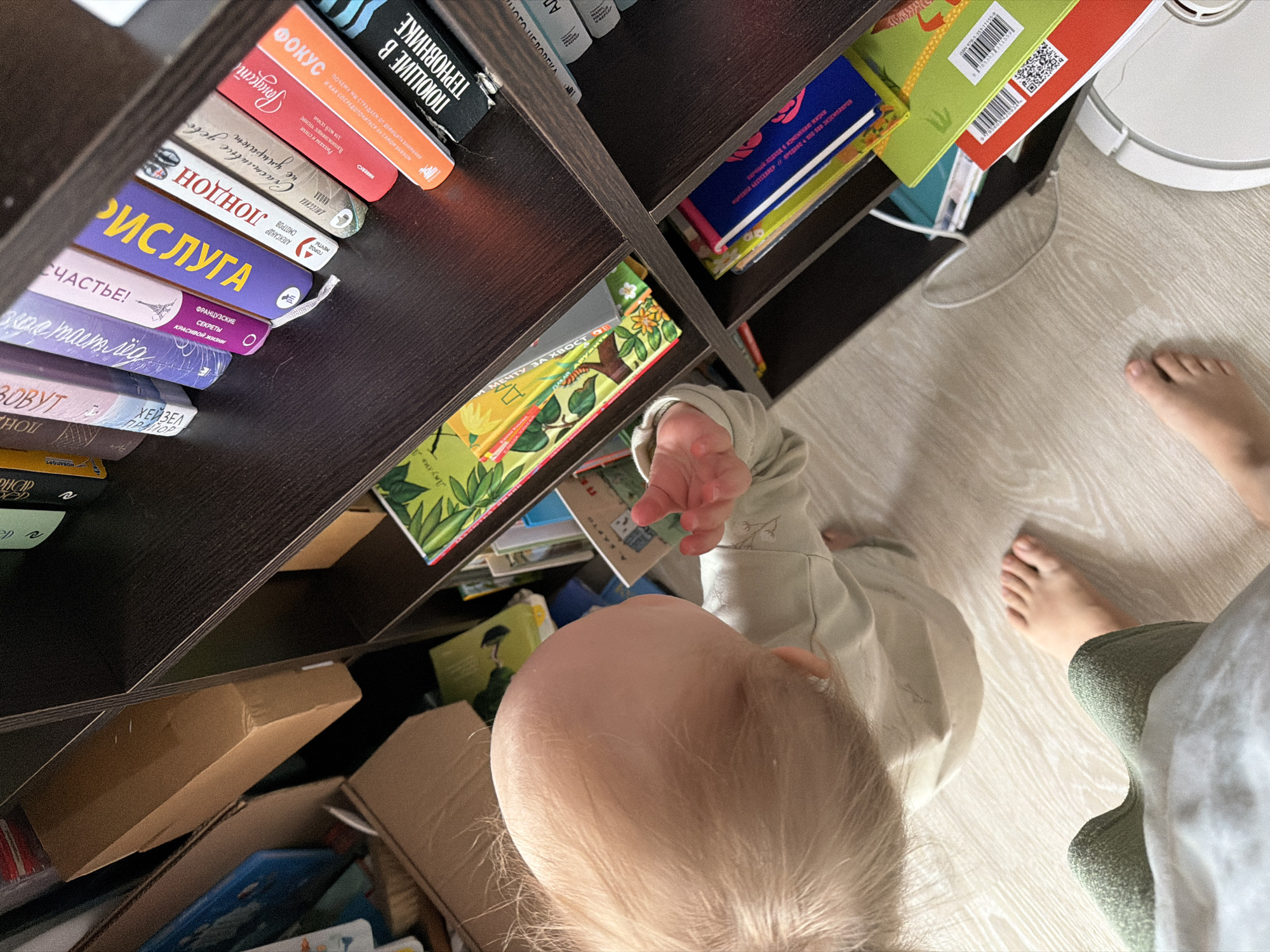 A top-down shot shows a child with blond hair reaching for books on a dark brown bookshelf. The books include Russian titles such as "Focus", "Risuluga", and "Happiness!". Some are children's books with colorful covers. Below the child, in the lowest shelf, are brown cardboard boxes and other books. Beside the bookshelf is a white robot vacuum cleaner, and on the light-colored wooden floor, are the feet of a person wearing green pants. It is impossible to determine the exact location of the scene in the city of Chaikovsky, Russia, without further information.
