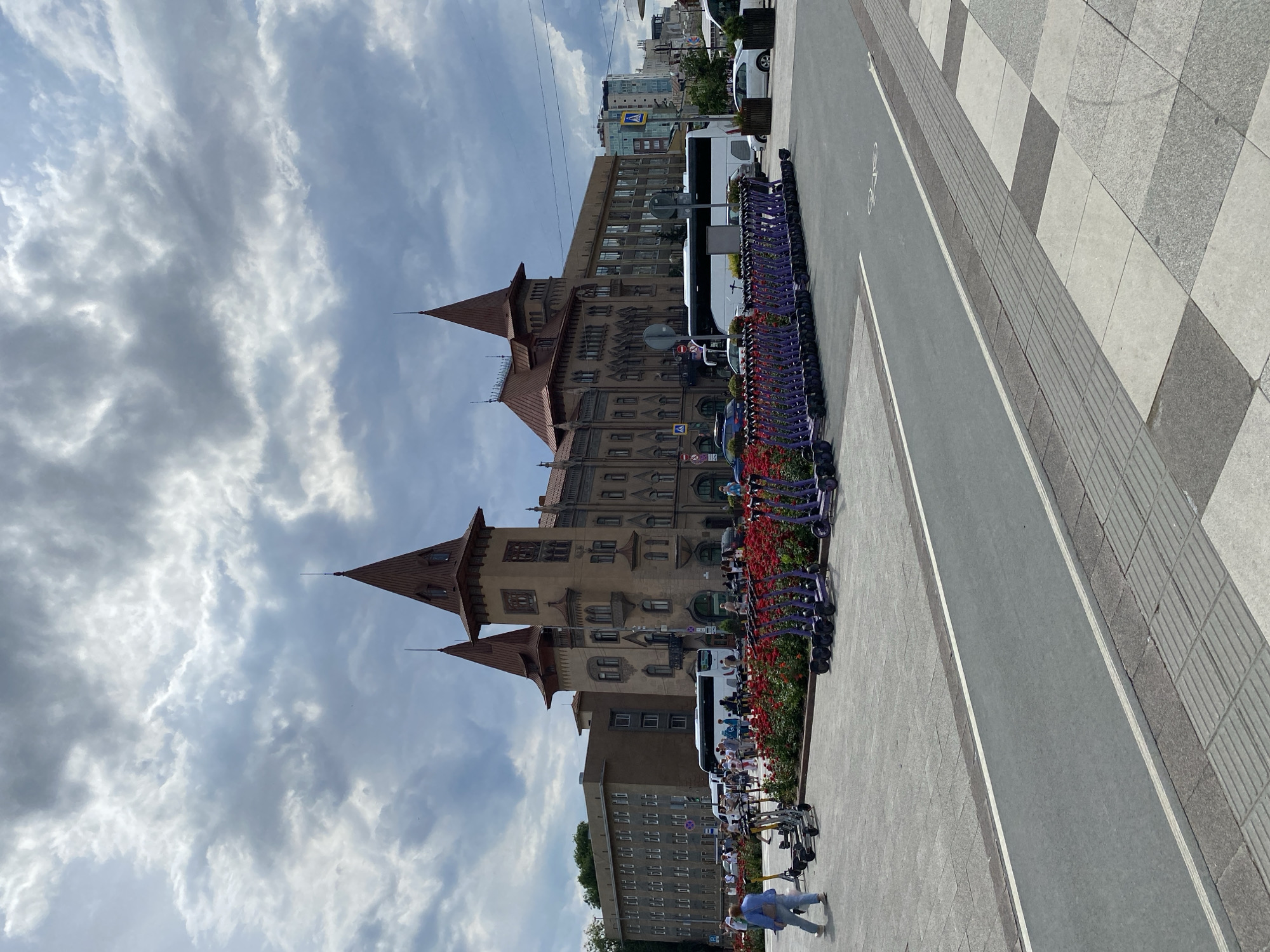 An exterior, eye-level, medium shot captures a public square in front of a large, ornate building with pointed turrets and a red roof. The building's facade is tan with dark windows and architectural detailing. In the foreground, a grey asphalt bike lane is marked with white lines. A row of parked purple electric scooters stretches across the middle ground, bordered by a vibrant red flowerbed. Several people are visible walking and standing around the scooters and in front of the building. A white bus is parked to the left of the building. The sky above is filled with dramatic, dark grey clouds, suggesting overcast weather. To the right of the main building, a taller, modern glass building is visible, along with street signs and traffic lights.