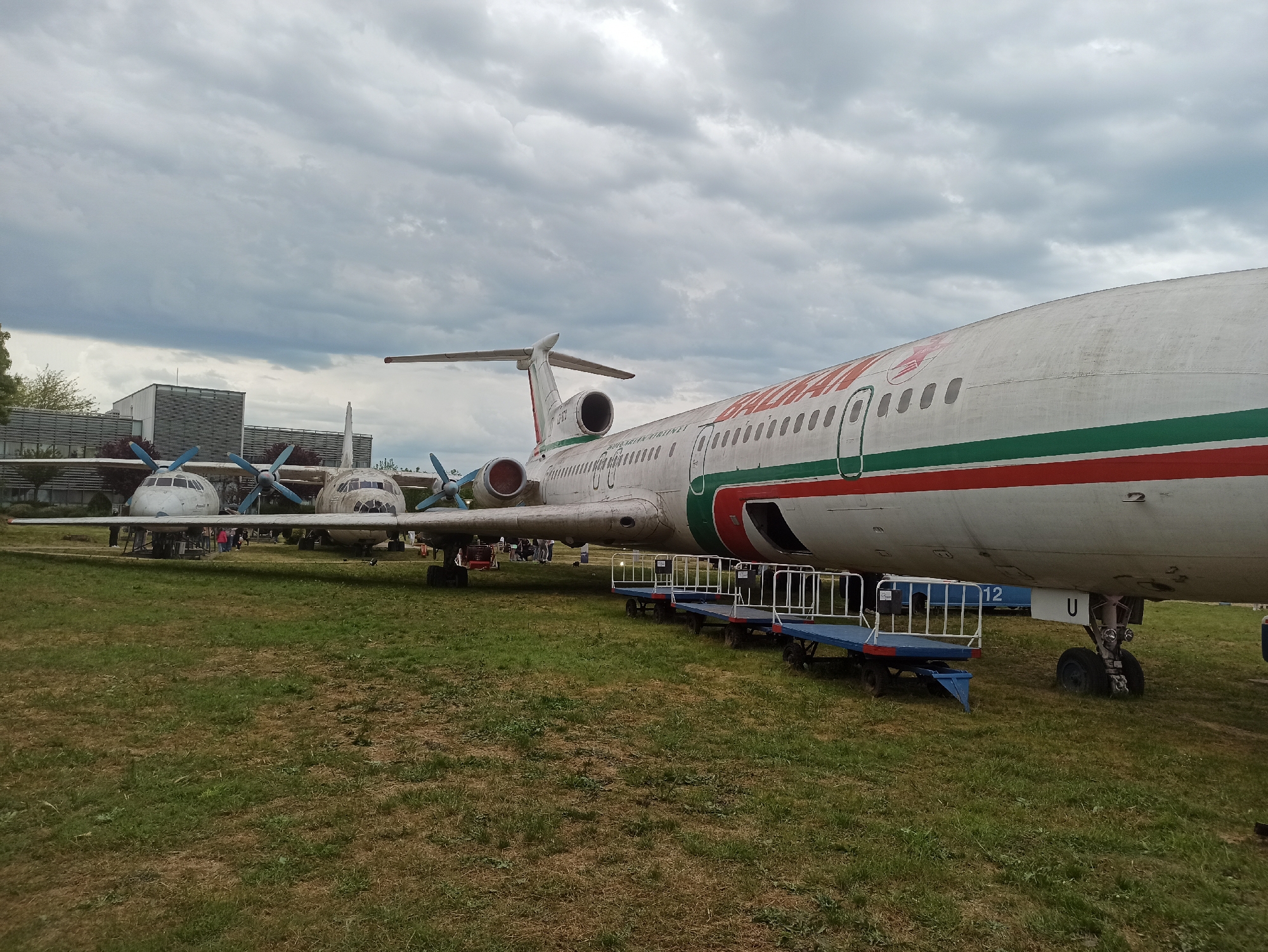 The image shows two aircraft on display in an open grassy area, likely part of an aviation museum or park. In the foreground is a Tupolev Tu-154, painted in a white livery with red and green stripes. The name "BALKAN" is visible on its fuselage. A baggage cart is parked beneath the aircraft. In the background are two turboprop aircraft with blue propellers. A building with a modern facade stands near the aircraft. The sky is overcast with heavy clouds. The location is Burgas, Bulgaria, inferred from the planes on display at the Burgas Aviation Museum.