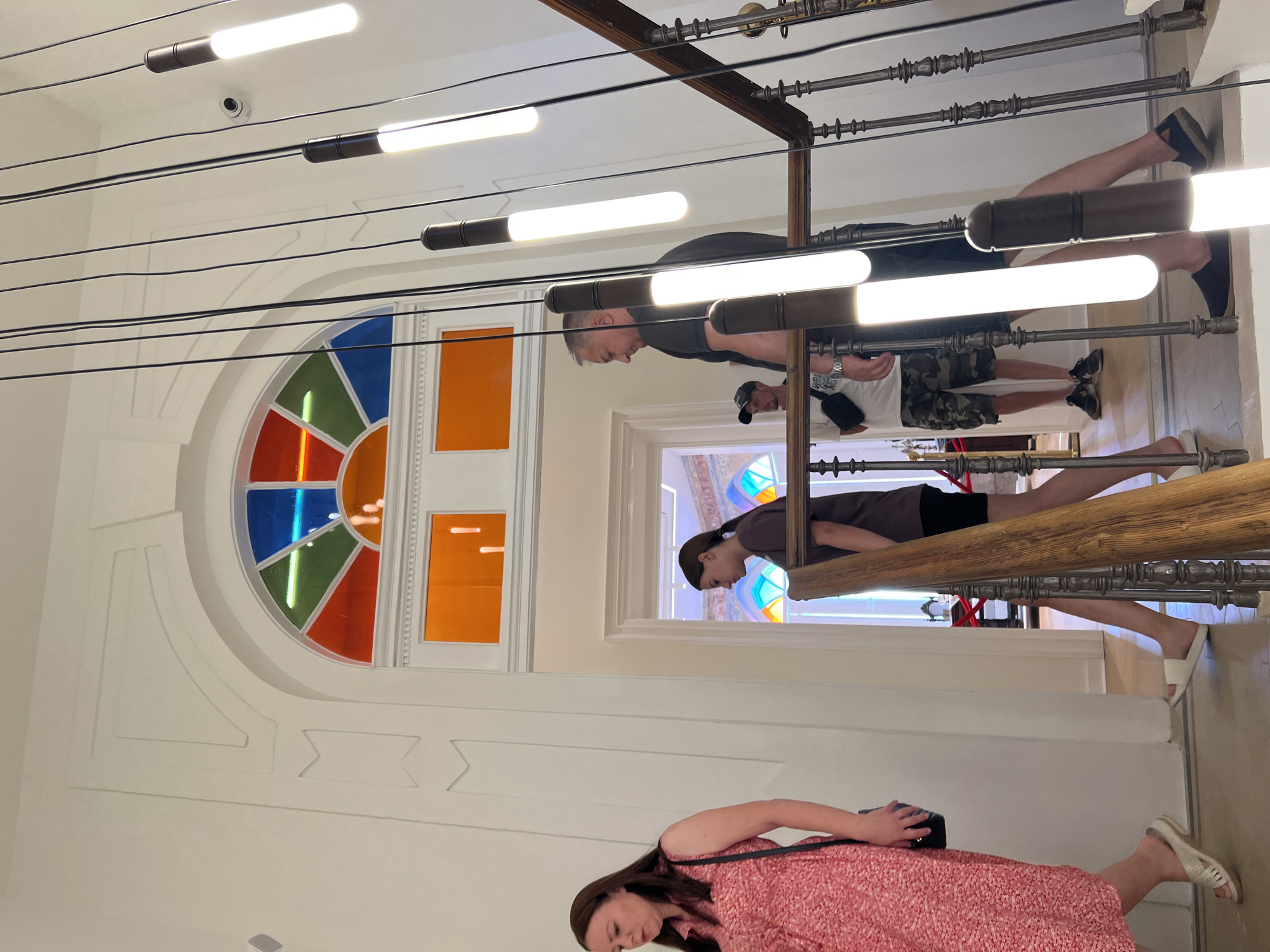 The image depicts an indoor staircase with several people ascending or descending. In the foreground, a woman in a red floral dress walks towards the left. Behind her and to the right, a young woman in a dark shirt and black shorts walks up the stairs. Further up the stairs, a man in a dark shirt and shorts is visible, and behind him, a man in a camouflage shirt and a baseball cap is also present. The staircase features ornate metal railings and a wooden handrail. Above the staircase, a large arched window is segmented into colorful panes of red, orange, green, and blue glass. Several modern, cylindrical pendant lights hang from the ceiling, illuminating the space. The architecture suggests a building with some historical or artistic significance. The context provided places this scene in the city of Bar, Montenegro.