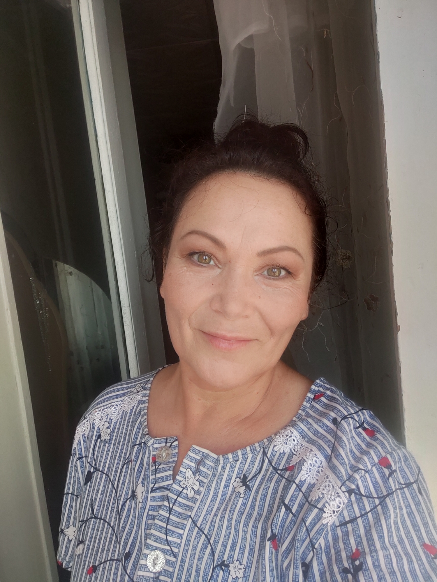 A medium shot shows a woman in her late 40s or early 50s taking a selfie in what appears to be an apartment in Oktyabrsky, Russia. She has dark brown hair pulled back into a messy bun, with loose strands framing her face. She is wearing a blue and white striped shirt with a floral pattern and lace detailing at the neckline. The woman has light skin, green eyes, and is wearing makeup. She is smiling slightly at the camera. Behind her, a window with white trim is visible, and sheer curtains hang to the right. The background is dark and out of focus, suggesting an indoor setting.