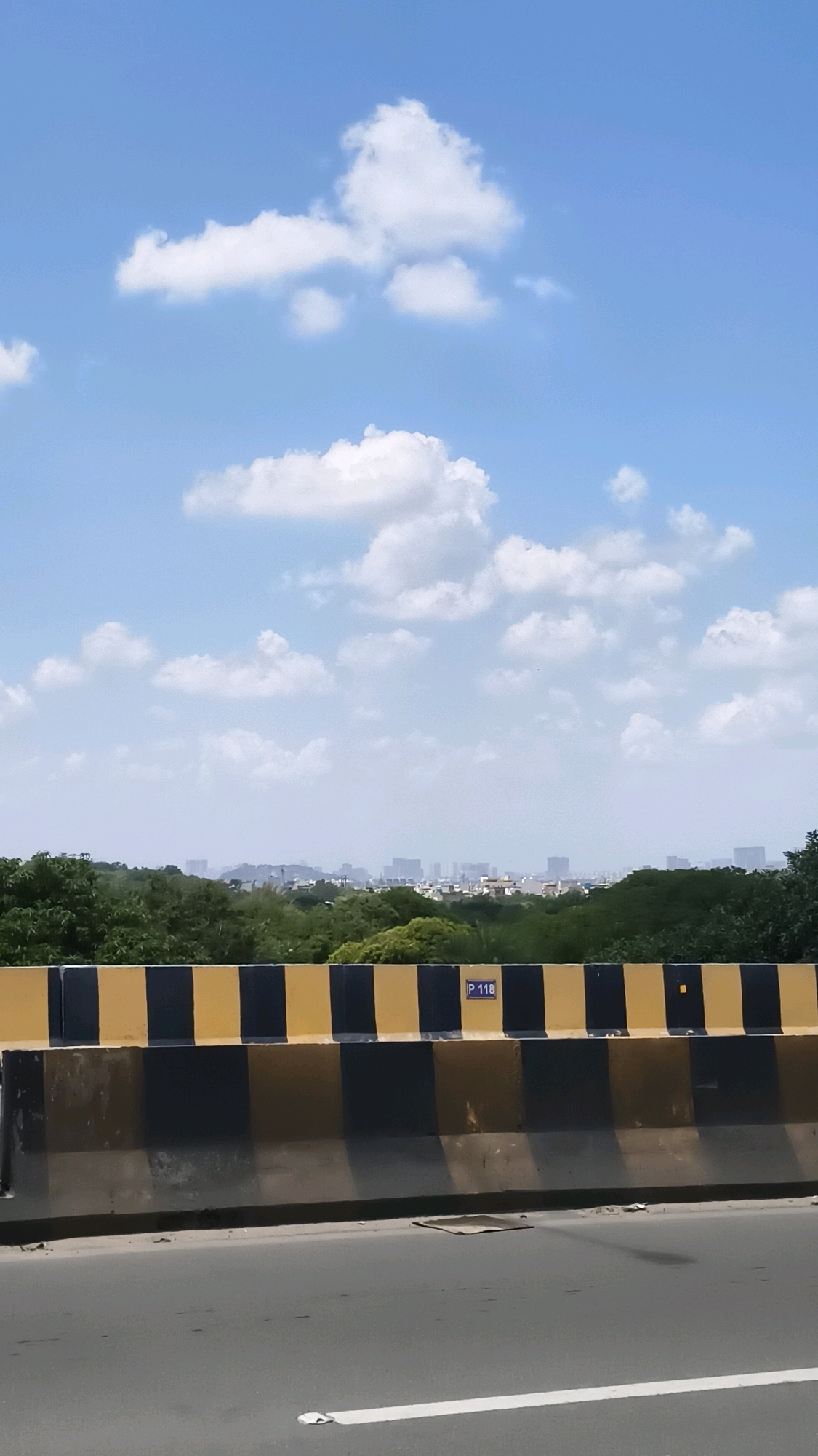 The image depicts a wide shot of a highway barrier in Hyderabad, India. The barrier is painted with alternating black and yellow stripes. Behind the barrier, lush green trees are visible, beyond which a distant cityscape with numerous buildings can be seen. The sky above is clear blue with scattered white clouds. The highway in the foreground is paved and has a white lane marker. A small sign with the text "P 118" is attached to the barrier. The scene suggests a bright, clear day.