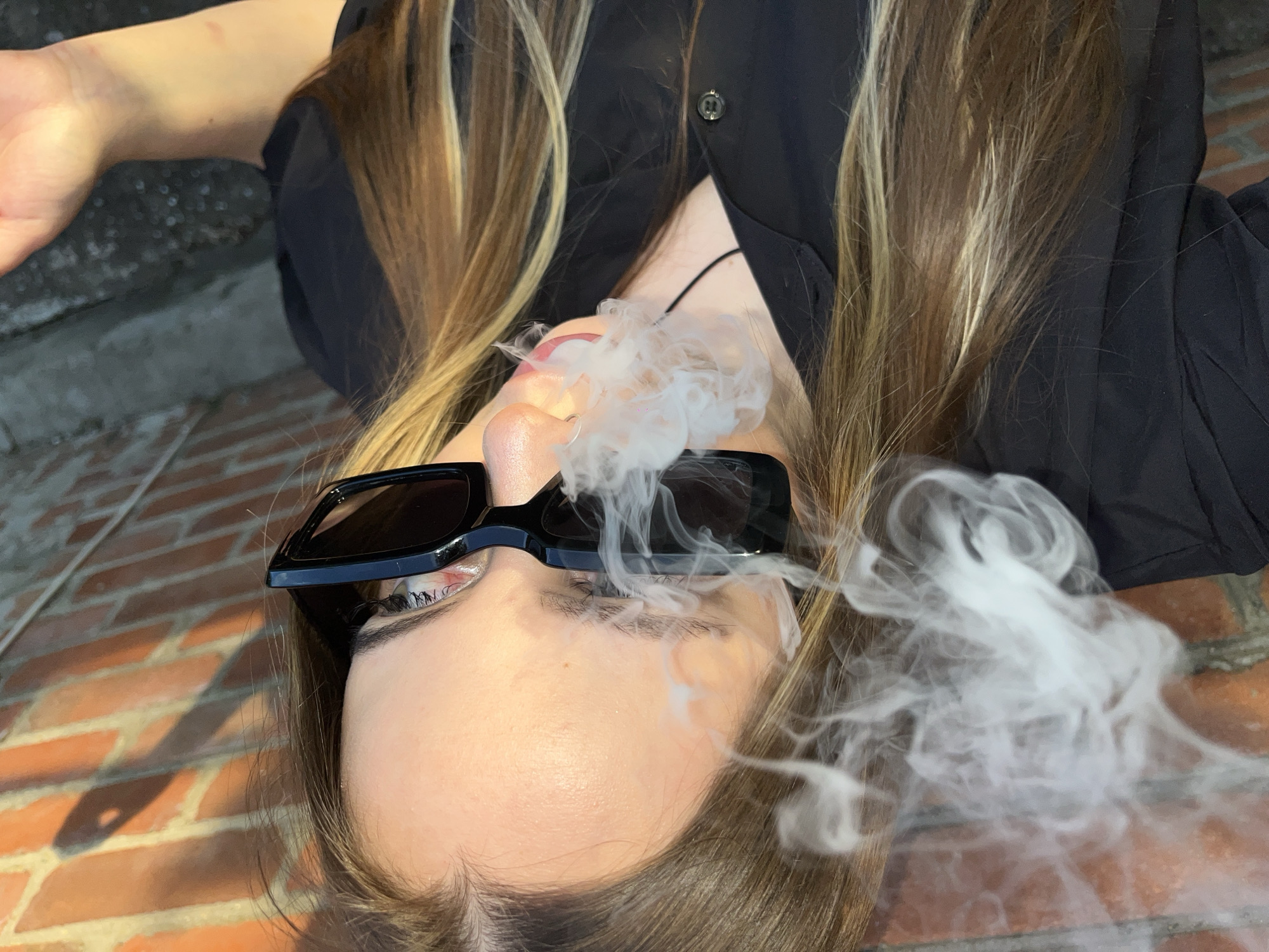An overhead, tilted shot captures a person, likely a young woman, lying outdoors in Novopokrovskaya, Russia, exhaling a large cloud of white smoke. The subject is viewed from the chest up, with her head oriented towards the bottom-center of the frame, looking upwards. She has long, light brown hair with prominent blonde highlights, which spreads around her head. She is dressed in a black collared shirt, unbuttoned at the top.
Her face is partially obscured by the billowing white smoke emanating from around her mouth and nostrils. She wears large, black, rectangular sunglasses that cover her eyes. A small silver hoop nose ring is visible in her left nostril. Her lips are slightly parted and appear to have pink lipstick. Her skin tone is light.
The background consists of two distinct surfaces: reddish-brown brickwork on the right and bottom, and a darker, textured, possibly concrete or asphalt surface in the upper left. Strong, elongated shadows are cast across the brickwork, suggesting bright daylight, possibly from a low sun angle like morning or late afternoon. A hand and wrist are partially visible in the upper left corner of the frame. The activity depicted is the exhalation or emission of a significant amount of smoke or vapor. No visible text is present in the image.