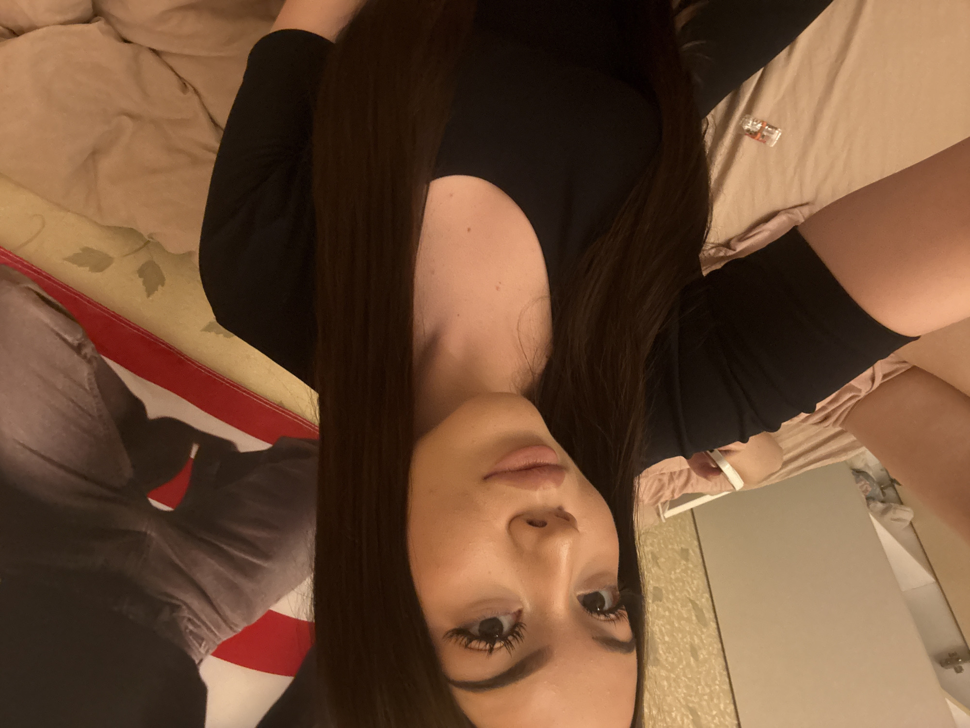 The image captures a young woman lying down and looking towards the camera, likely taking a selfie. She has long, dark brown hair that cascades around her, fair skin, and dark eyes with prominent eyelashes and defined eyebrows. Her lips are a soft, light pink. She is wearing a black long-sleeved top or dress with a scoop neckline. A white wristband is visible on her right wrist.
The setting appears to be an indoor space, likely a bedroom, with light beige or tan bedding. To her left, there's a segment of fabric featuring red and white stripes alongside a dark blue or grey area, resembling a flag pattern (potentially an American flag, or part of a blanket/clothing item). A small, clear plastic object, possibly an electrical connector or a similar small item, rests on the bed near her right arm. The lower right section of the image shows reflective surfaces, possibly a mirror or polished furniture, suggesting additional room elements. The lighting in the room is somewhat dim, creating an intimate atmosphere. The location is Naberezhnye Chelny, Russia. No visible text is present.