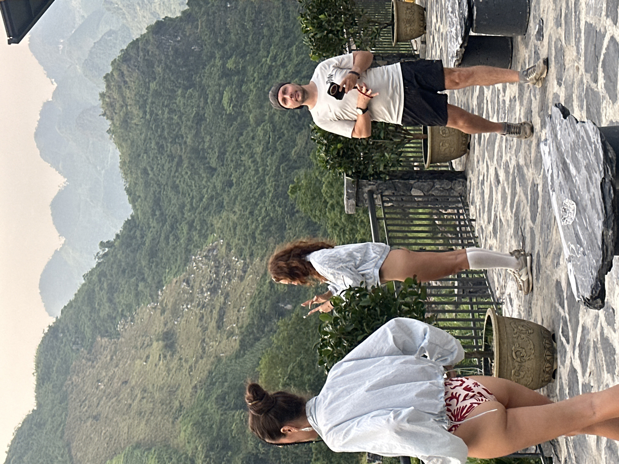 The image shows a group of three people on a stone patio overlooking a mountainous landscape. The backdrop appears to be the verdant hills of Du Già Commune, Vietnam. A man is standing on the right, wearing a white t-shirt, black shorts, sneakers, and a beanie. He holds a black object in his hand. Two women are in the foreground, both wearing light blue shirts. One woman is wearing red and white printed bottoms. The other woman is wearing white knee-high socks and sneakers. The people are standing near potted plants and a wrought-iron railing.