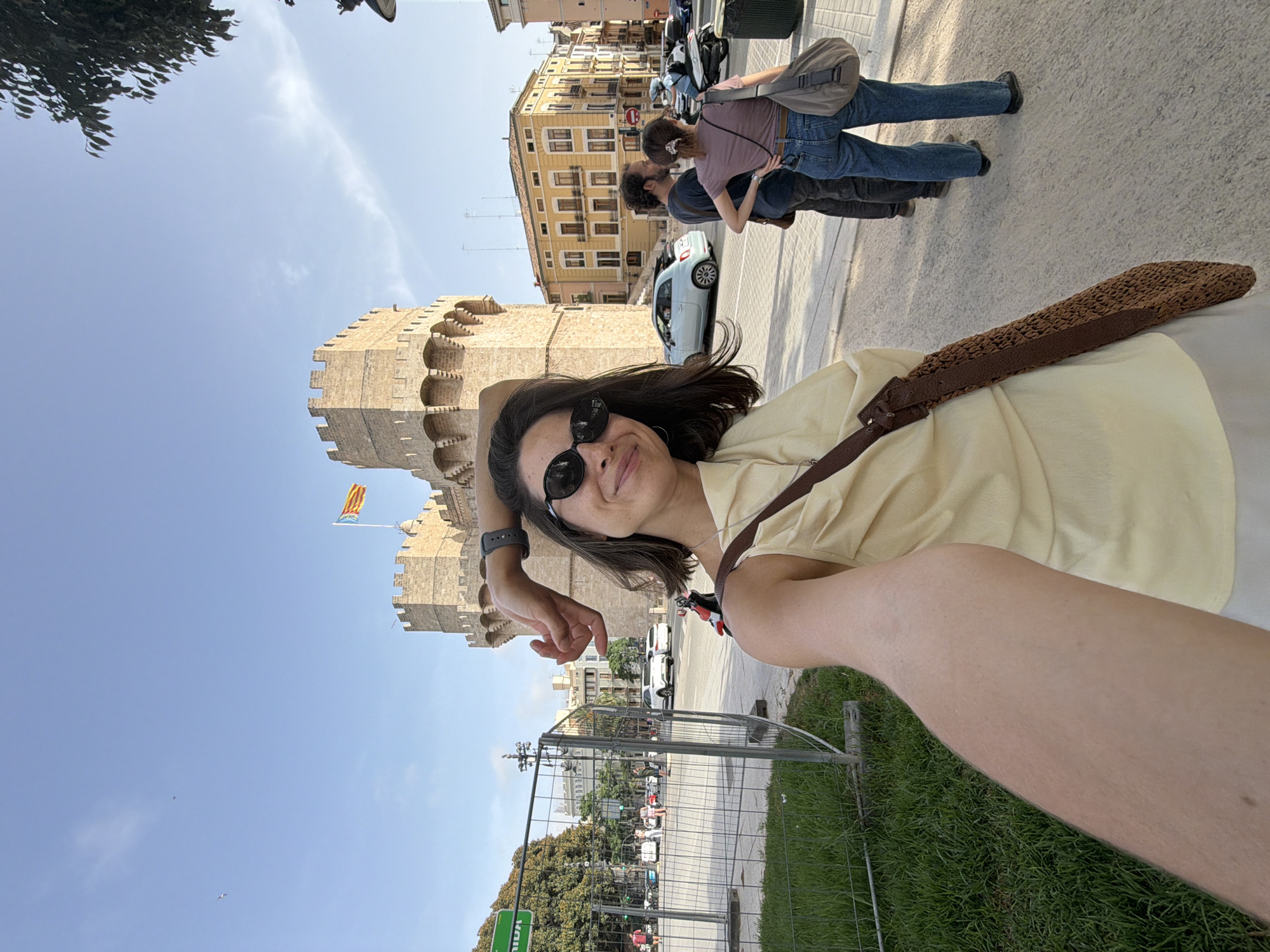 A woman takes a selfie in front of a stone tower in a city. She is wearing sunglasses and a yellow dress, with a brown bag slung over her shoulder. In the background, two people are standing on a sidewalk, and there are buildings and cars visible. The sky is blue with a few clouds.