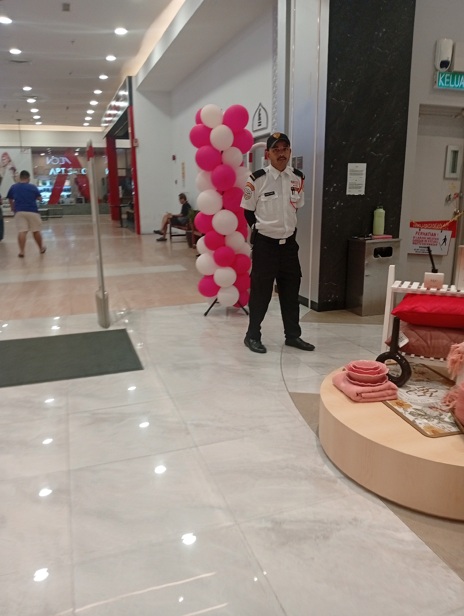 The image captures an indoor scene, likely within a shopping mall in Kuala Lumpur, Malaysia. A security guard, dressed in a white shirt with a dark tie and black pants, stands near a display of pink and white balloons. He is positioned slightly to the right of the frame's vertical center. To the left of the guard, a person in a blue t-shirt and light-colored shorts is walking away from the camera, with another person partially visible in the background. Further in the background, a person sits on a bench.
The environment features a polished floor, illuminated by overhead lighting. A sign with text in a foreign language is visible on the wall behind the security guard, and a trash receptacle is nearby. A white shelving unit with pink cushions is on the right side of the frame, and a round display table with stacked pink bowls and towels is in the foreground. The overall lighting suggests it is daytime or well-lit indoor environment.