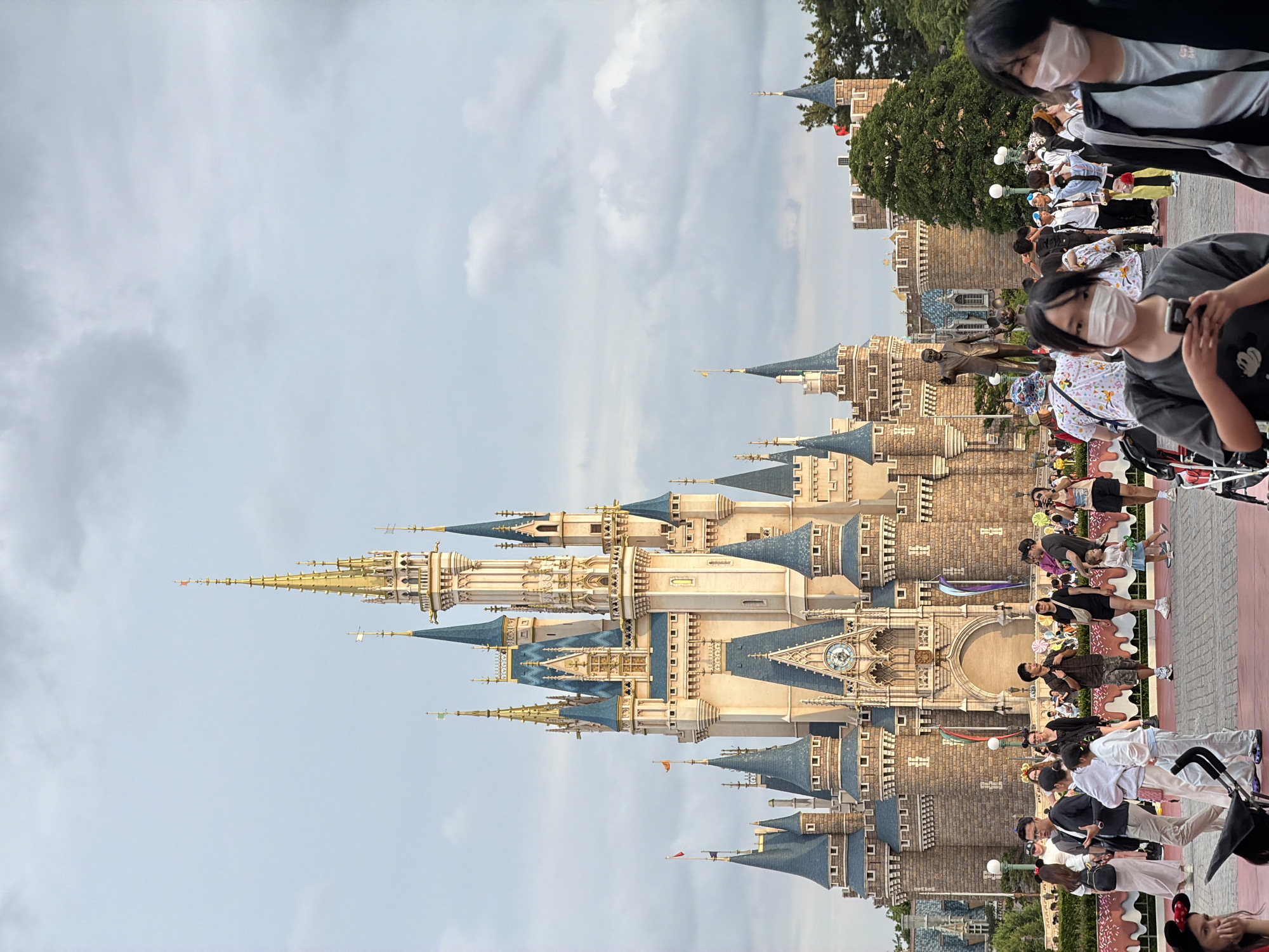 This is a full shot, eye-level, vertical photograph of Cinderella Castle at Tokyo Disneyland in Urayasu, Japan. The sky is overcast with scattered clouds. The castle, made of light brown stone with blue turrets and gold spires, dominates the background. A statue of Walt Disney and Mickey Mouse stands in front of the castle. In the foreground, a crowd of people, some wearing face masks, are walking and interacting. The ground is paved with red bricks.