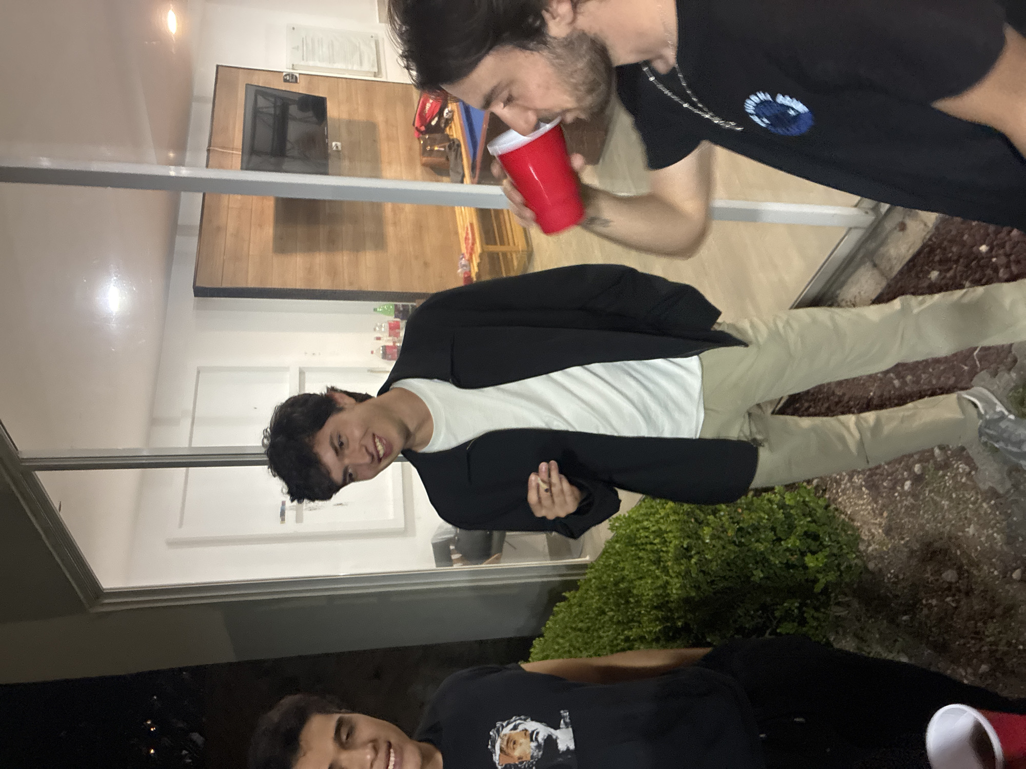 Three individuals are visible, likely outdoors adjacent to a building. One person, facing the camera, wears a white t-shirt under a black blazer, and tan pants, holding what appears to be a cigarette. A second person is drinking from a red plastic cup. The third person is wearing a black t-shirt with an image on it, black pants, and is near another red plastic cup. The building's interior is visible through windows. The exterior has a bush and ground cover. The location could be in Huixquilucan de Degollado, Mexico, based on potential identifying factors outside of the provided image, but cannot be determined from the image itself.