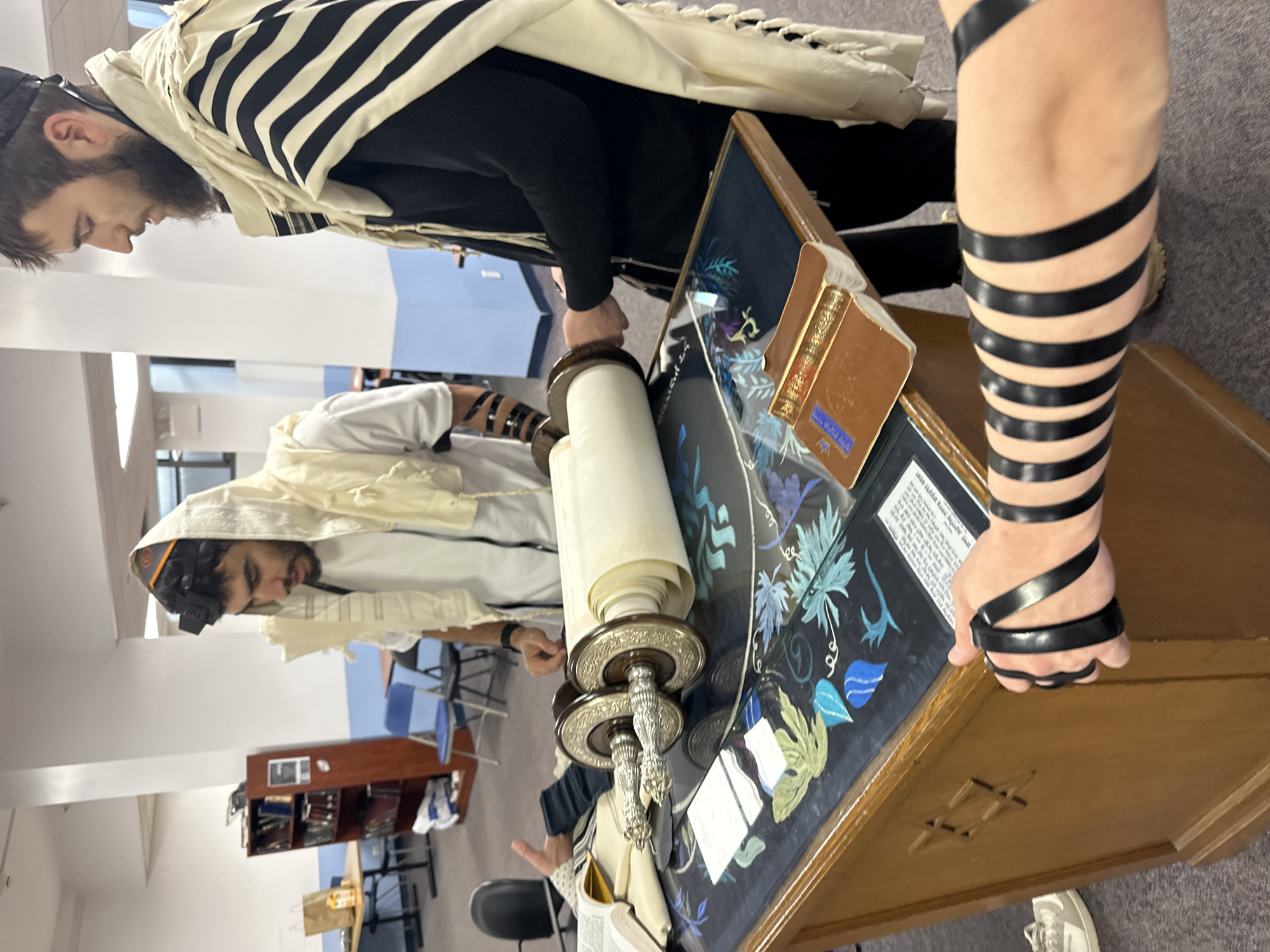 Two men are engaged in a Jewish religious ritual, likely a prayer service, in an indoor setting that appears to be a synagogue or study hall. The image is taken from a slightly elevated perspective, looking down at the participants and the objects on a wooden lectern.
In the foreground, a man's right arm and hand are visible, wrapped in black leather phylactery (tefillin) straps. He is positioned next to a decorated wooden lectern, which features a carved Star of David on its side and a surface adorned with blue, green, and purple floral motifs.
At the center of the scene, another bearded man is leaning over the lectern, preparing or interacting with a large Torah scroll. He wears a white and black striped tallit (prayer shawl) draped over his shoulders and head, and has a black phylactery box (tefillin shel rosh) on his forehead. His hands are near the silver, ornate handles of the Torah scroll.
To the left and further back, a third bearded man is partially visible, also wearing a tallit and kippah. In the background, typical interior elements like tables, chairs, bookshelves filled with books, and fluorescent lighting are visible. The walls are light-colored, with a section of light blue.
On the lectern, beside the Torah scroll, there is a small brown book with gold lettering, likely a prayer book or holy text, and a small white placard with illegible English text. The men's attire and the ritual objects clearly indicate a Jewish religious context.