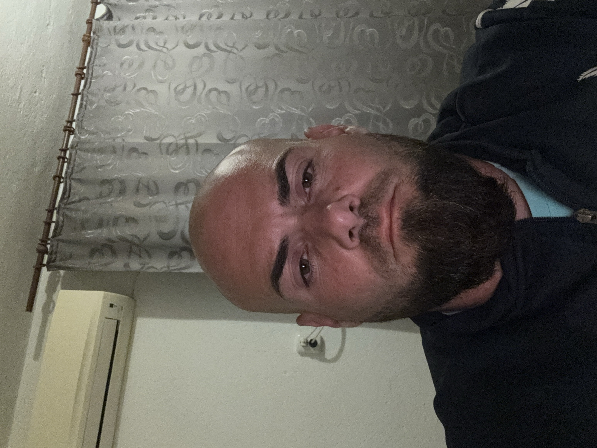 A selfie of a bald man with a beard, wearing a dark colored, possibly navy blue, sweatshirt over a light blue shirt. The background shows a wall with an air conditioning unit, a light switch, and a window covered with a patterned curtain. The image does not contain location information to confirm the presence in Sarnevo, Bulgaria.