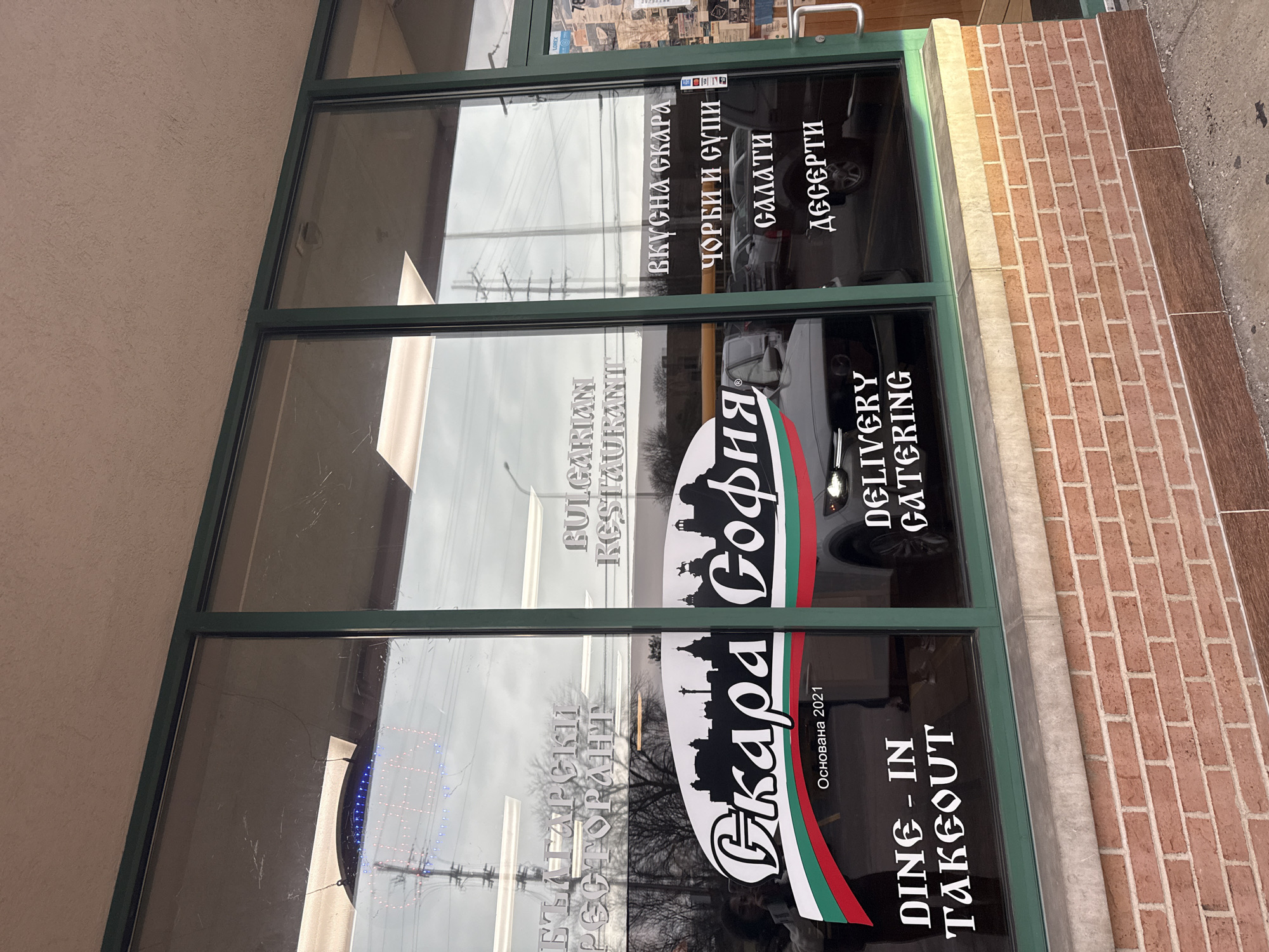 The image shows the storefront of a Bulgarian restaurant in Des Plaines, Illinois, USA. The restaurant's name is "Skara Sofia", indicating that it specializes in grilled dishes ("skara"). The logo incorporates the skyline of Sofia, Bulgaria and the Bulgarian flag. The signage includes the words "Bulgarian Restaurant" in both English and Bulgarian ("БЪЛГАРСКИ РЕСТОРАНТ"). It also specifies "Dine-In/Takeout" and "Delivery/Catering". The menu highlights "delicious grilled dishes" ("ВКУСНА СКАРА"), "soups" ("ЧОРБИ И СУПИ"), "salads" ("САЛАТИ") and "desserts" ("ДЕСЕРТИ").