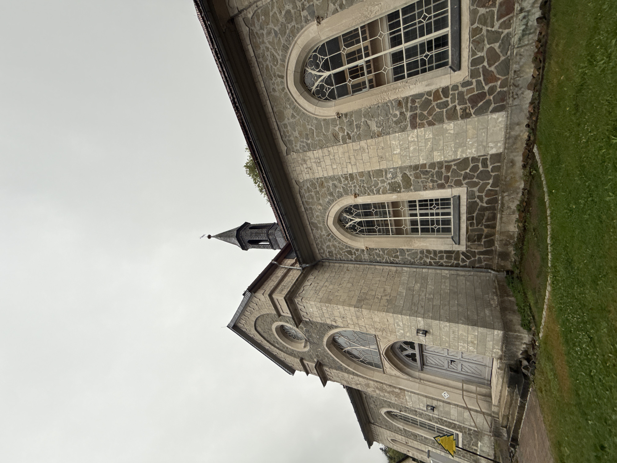 This is a ground-level, wide-angle photograph of the exterior of the historic St. Nicholas' Church in Walkenried, Germany, captured on an overcast day. The camera is positioned low, looking up at the building, emphasizing its imposing architecture.
The church is constructed primarily from large, roughly hewn stones, giving it a rustic and weathered appearance. Interspersed with the stone are sections of lighter, rectangular blocks, creating a patterned effect on the facade. The building features Gothic architectural elements, most notably its tall, arched windows with intricate white mullions. One prominent window on the right side of the image is particularly large and detailed. Another arched window is visible above the main entrance, along with a circular window set higher up on the gable.
The entrance is marked by a light gray, double-door with recessed panels and a rounded archway above. A set of stone steps leads up to the door, with a wooden handrail on the left. A yellow triangular warning sign is visible on a pole to the left of the entrance.
A slender, dark steeple with arched openings and a pointed roof rises from behind the main structure. The roof of the church itself is dark and appears to be tiled.
The foreground consists of a grassy lawn, which appears somewhat unkempt, with patches of bare soil and scattered stones. A low stone border separates the grass from the church's foundation. The sky is a uniform gray, contributing to the muted and somewhat somber atmosphere of the scene. No people are visible in the photograph.