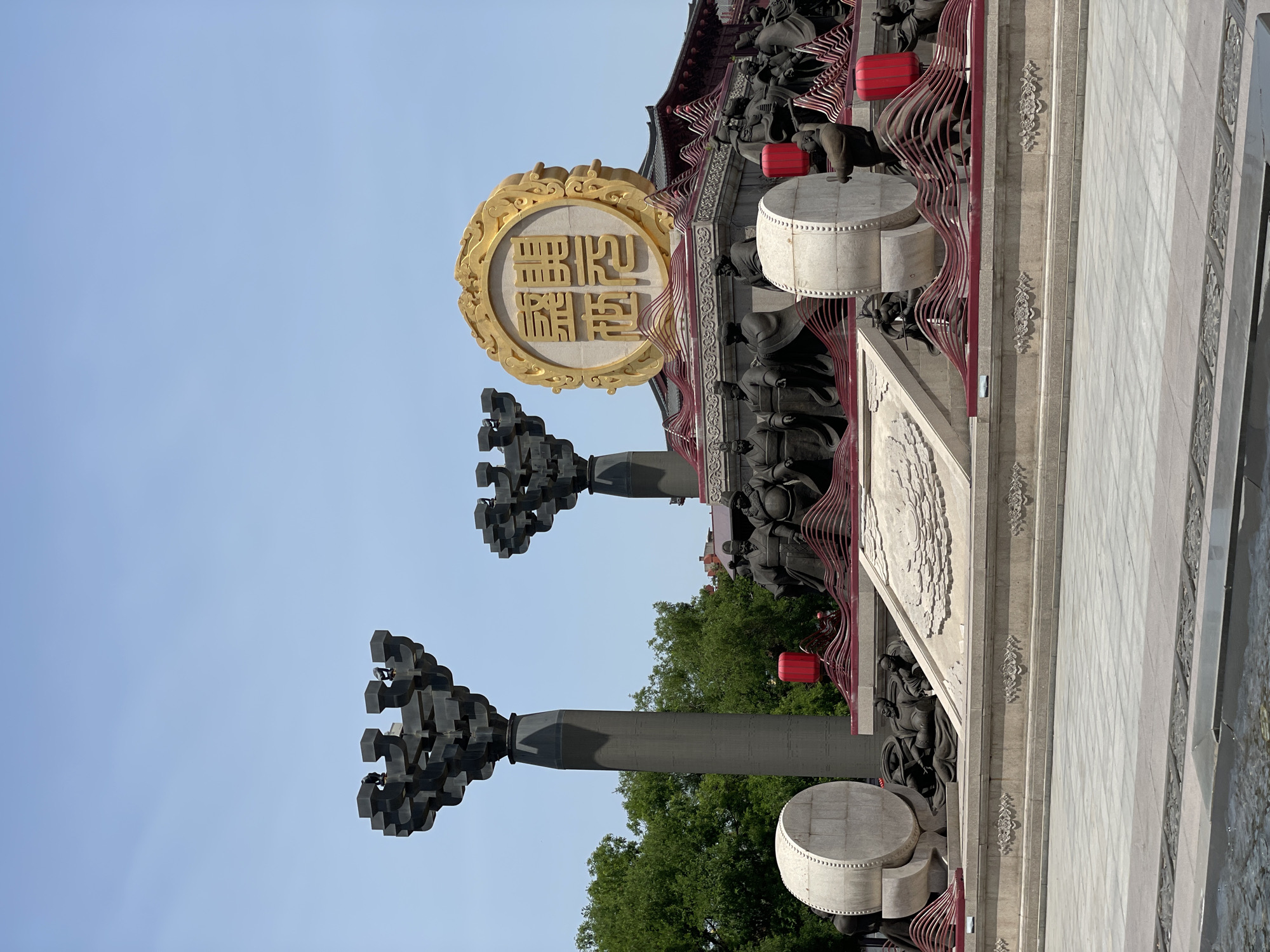 This image shows a portion of a building or monument in the Yanta District of Xi'an, China. Prominently displayed is a large, round, gold-colored emblem with Chinese characters, mounted on the building's facade. Below the emblem are two large, gray-colored pillars topped with multiple dark, cuboid-shaped elements. Red lanterns are also visible. The architecture includes detailed carvings and statues of figures. Trees are also visible in the background.