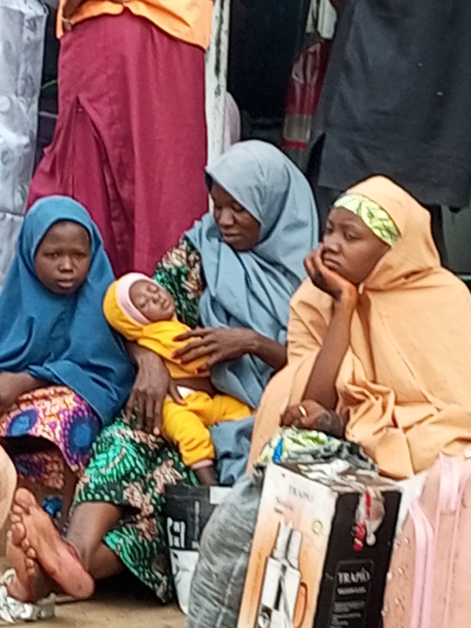 The image depicts a group of individuals, likely at a marketplace or gathering point in Gombe, Nigeria. In the foreground, a woman wearing a light orange hijab with a green patterned headscarf is seated, resting her chin on her hand and looking to the side. She is holding a baby swaddled in a bright yellow garment, whose face is not clearly visible. Beside her, to the left, a younger person wearing a blue hijab and a patterned skirt is also seated. Another woman, wearing a grey hijab, is seated behind the baby, with her arms around it. The background shows parts of other people and a large red garment, possibly a wrapper or skirt, draped over something. Various objects are scattered around, including a partially opened cardboard box with the brand name "TRAPO" visible, and parts of luggage, one of which is pink. The ground appears to be dusty or sandy. The overall scene suggests a moment of rest or waiting in a public, possibly crowded, setting.