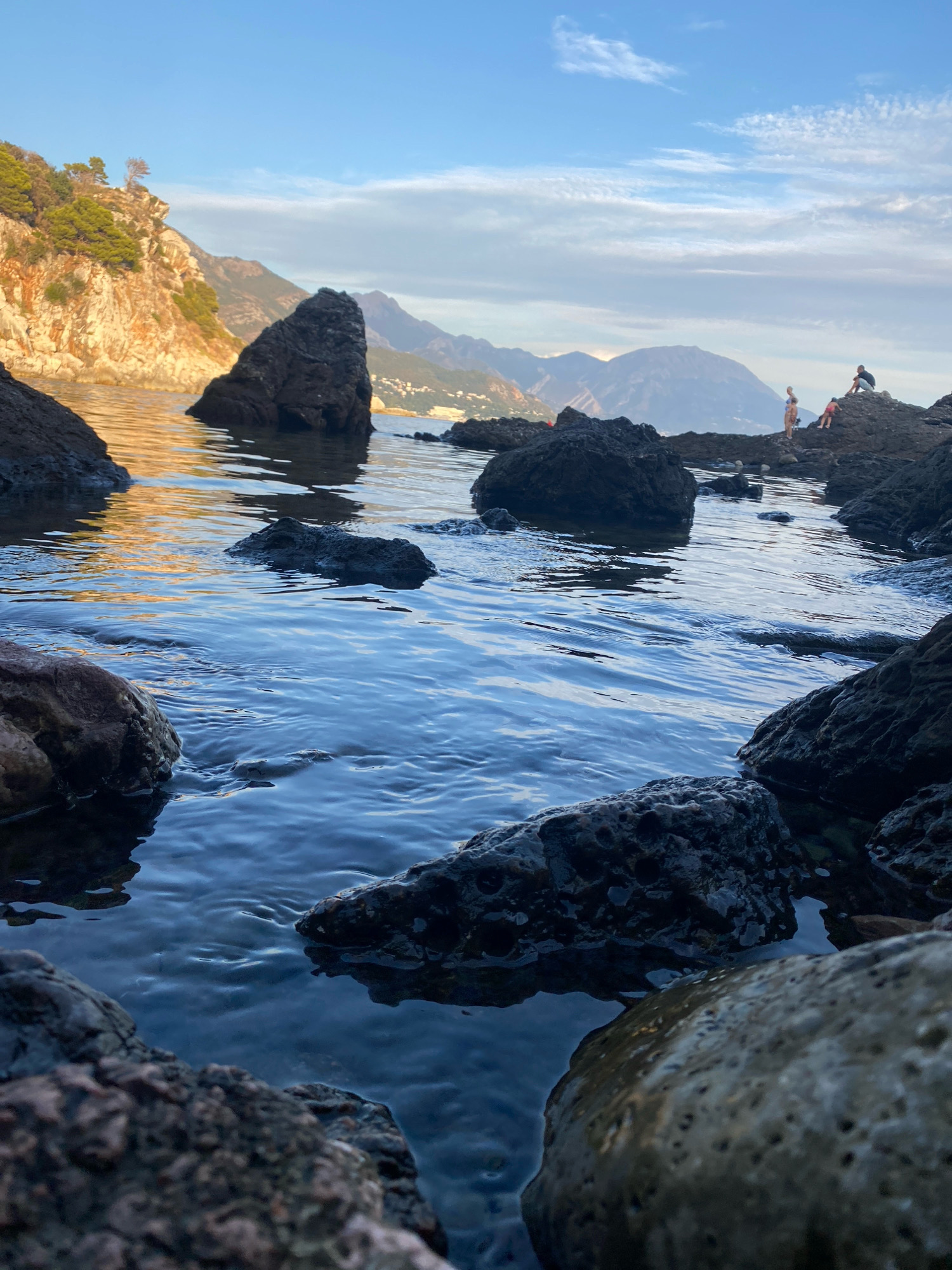 This is a low-angle, close-up shot of the rocky coastline at Sutomore, Montenegro, with the Adriatic Sea in the foreground. The water is a deep blue, reflecting the sky and surrounding rocks. Several dark, jagged rocks jut out of the water, with some partially submerged. In the background, a rugged, tree-covered cliff face is illuminated by the setting sun, casting a warm golden glow on the water. Further in the distance, hazy mountains rise, with a small town nestled on their slopes. On a distant rocky outcrop, two individuals, a woman and a child, are visible, along with another person sitting on the rocks. The sky is a clear blue with scattered, wispy clouds.