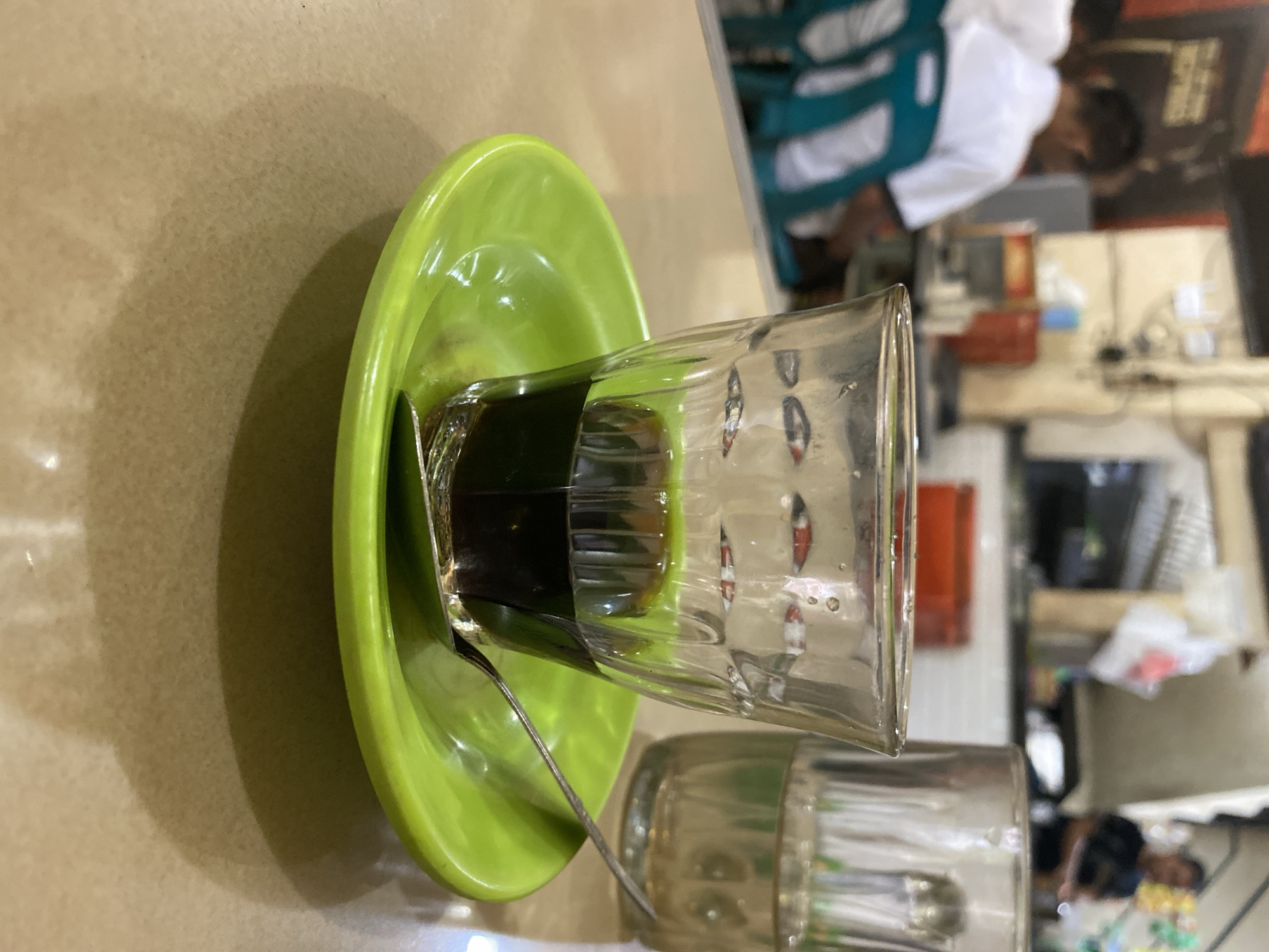 A close-up shot focuses on a clear glass containing a dark liquid, likely coffee, placed on a bright green saucer. A small silver spoon rests within the saucer, partially under the glass. In the background, out of focus, other patrons are visible sitting at tables, and there appears to be a serving counter or kitchen area. The overall setting suggests a casual eatery or coffee shop, possibly in Lhokseumawe, Indonesia, given the context. The lighting is natural and diffused, creating a warm and inviting atmosphere.