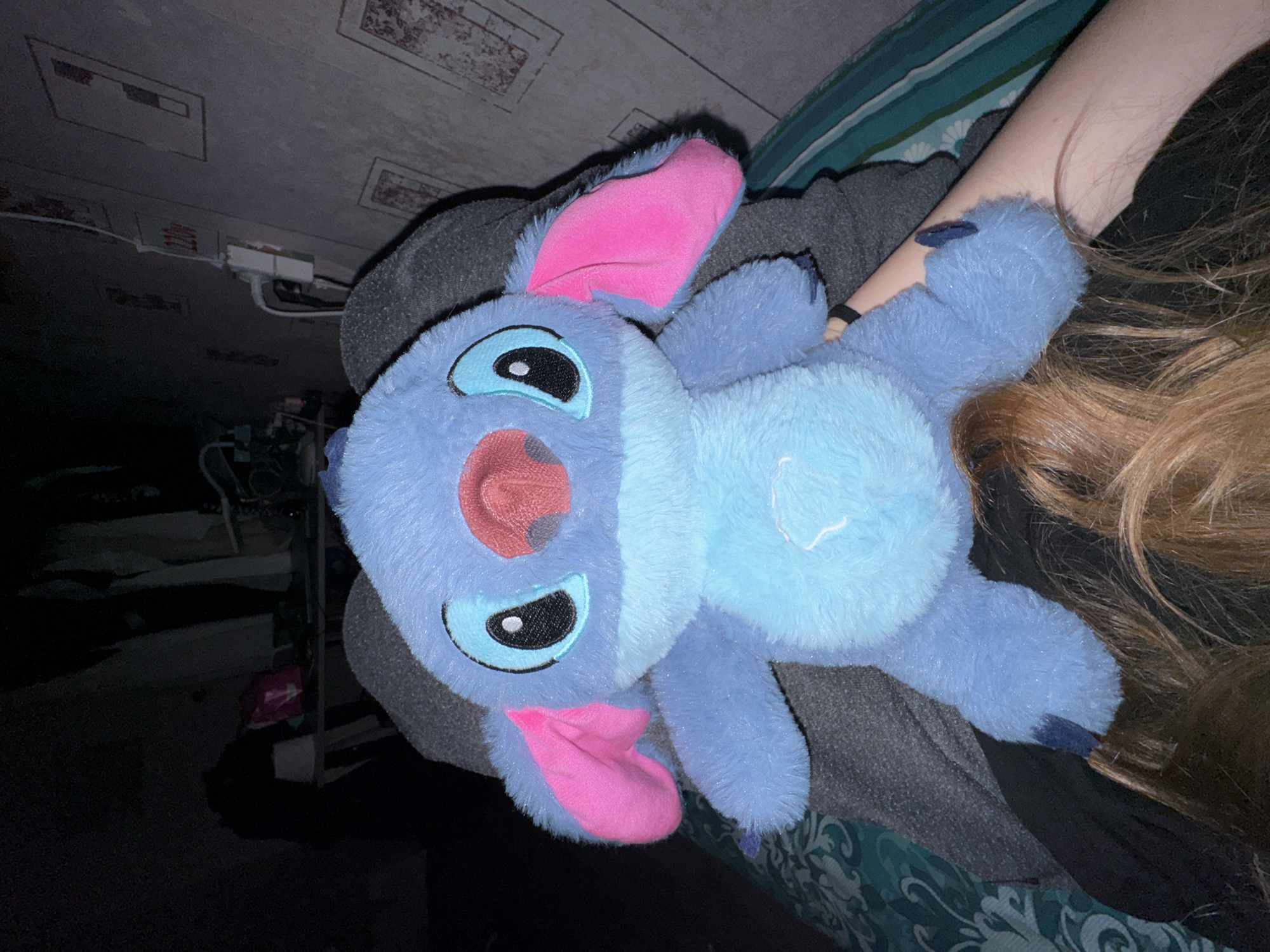 The image, taken in Perm, Russia, depicts a person holding a blue plush toy of the Disney character Stitch in a dimly lit indoor setting. The central subject is the plush toy, which is light blue with a lighter blue belly patch, vibrant pink inner ears, a pink nose, and black eyes with light blue outlines. It is held by a person whose fair-skinned right arm and long, light brown hair are visible on the right side of the frame. The person appears to be wearing dark clothing, possibly a grey or black hoodie, which is partially visible around the toy.
The activity taking place is a person holding or embracing the plush toy, suggesting a moment of comfort or affection.
Notable details include the time of day, which appears to be nighttime or very late due to the extremely dim ambient lighting in the room. A direct light source brightly illuminates the plush toy and the visible parts of the person, contrasting sharply with the dark background. The background reveals a room with a patterned ceiling featuring rectangular panels, some of which have faint, unidentifiable designs. A white electrical box with connected wires is mounted on the wall or ceiling. Other objects in the background are obscured by darkness but appear to be typical indoor furnishings. A green and white patterned fabric, possibly bedding, is visible on the far right. Some faint, unreadable red text is discernible on the white electrical component in the background.