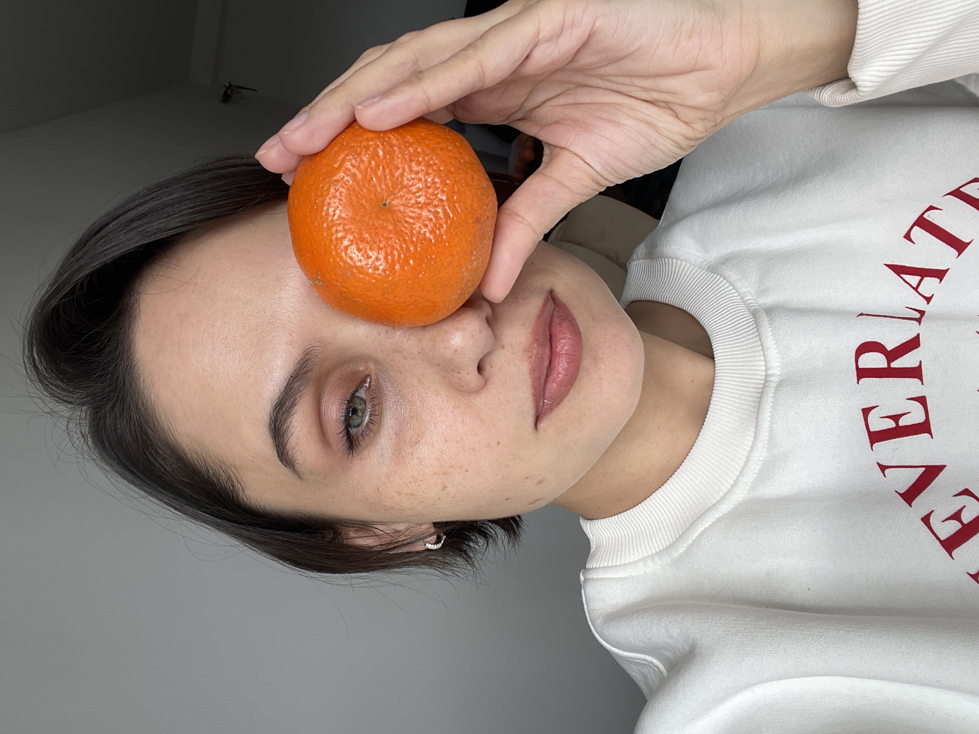 A young adult female with short dark hair is holding a tangerine over her right eye. The woman has fair skin, light-colored eyes, and some freckles. She wears a white sweatshirt with red lettering. A small earring is visible in her left ear. The background appears to be a plain gray wall and a light-colored ceiling. No information in the image indicates it was taken in Izhevsk, Russia.