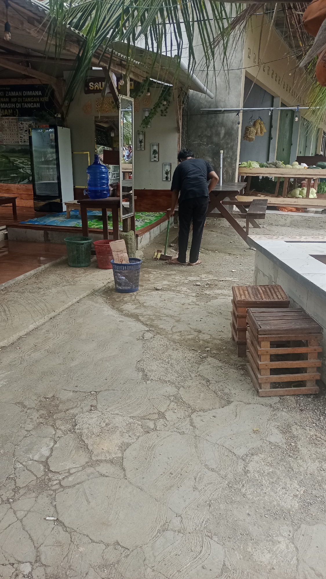The image depicts an outdoor area with a concrete ground surface that is cracked and has a textured pattern. In the foreground, there are three plastic trash cans, two of which are green and red, and one is blue. A person wearing a black t-shirt and dark pants is standing on the concrete and sweeping the ground with a broom. The person is facing away from the camera. Behind the person, there is a wooden picnic table and benches. In the background, there is a building with a thatched roof and a display of fruits and vegetables on a wooden table. There is also a large blue water jug on a stand. Lush green palm fronds hang from overhead. The scene appears to be a casual outdoor dining or market area. The location is identified as Cibeber, Indonesia.