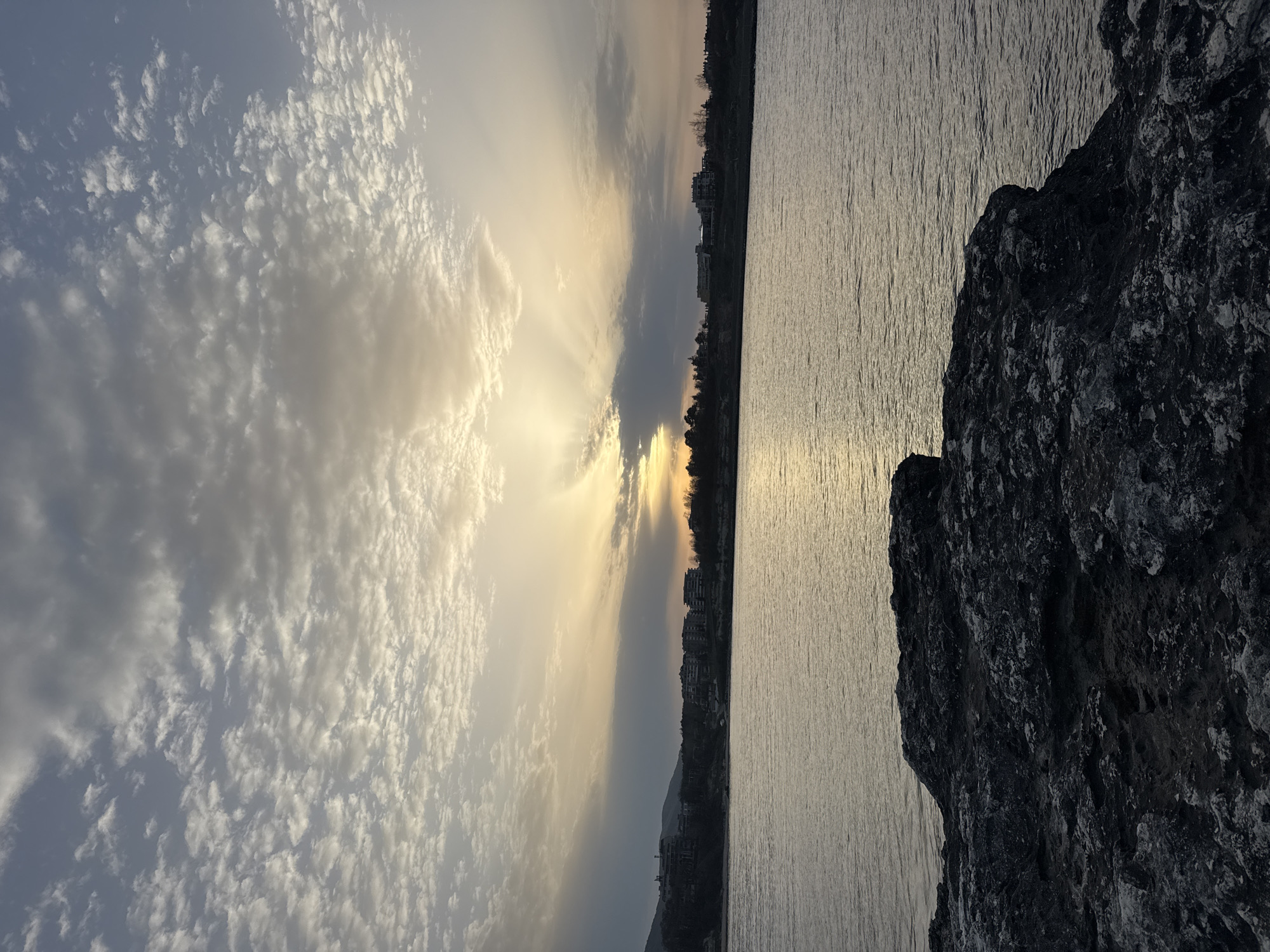 The image captures a coastal scene at dusk in Tsarevo, Bulgaria. The sky is dominated by altocumulus clouds, with sun rays visible around the horizon. The buildings are visible on the coastline across a body of water. A rocky outcrop occupies the right foreground. The water surface appears calm with reflections of the sky.