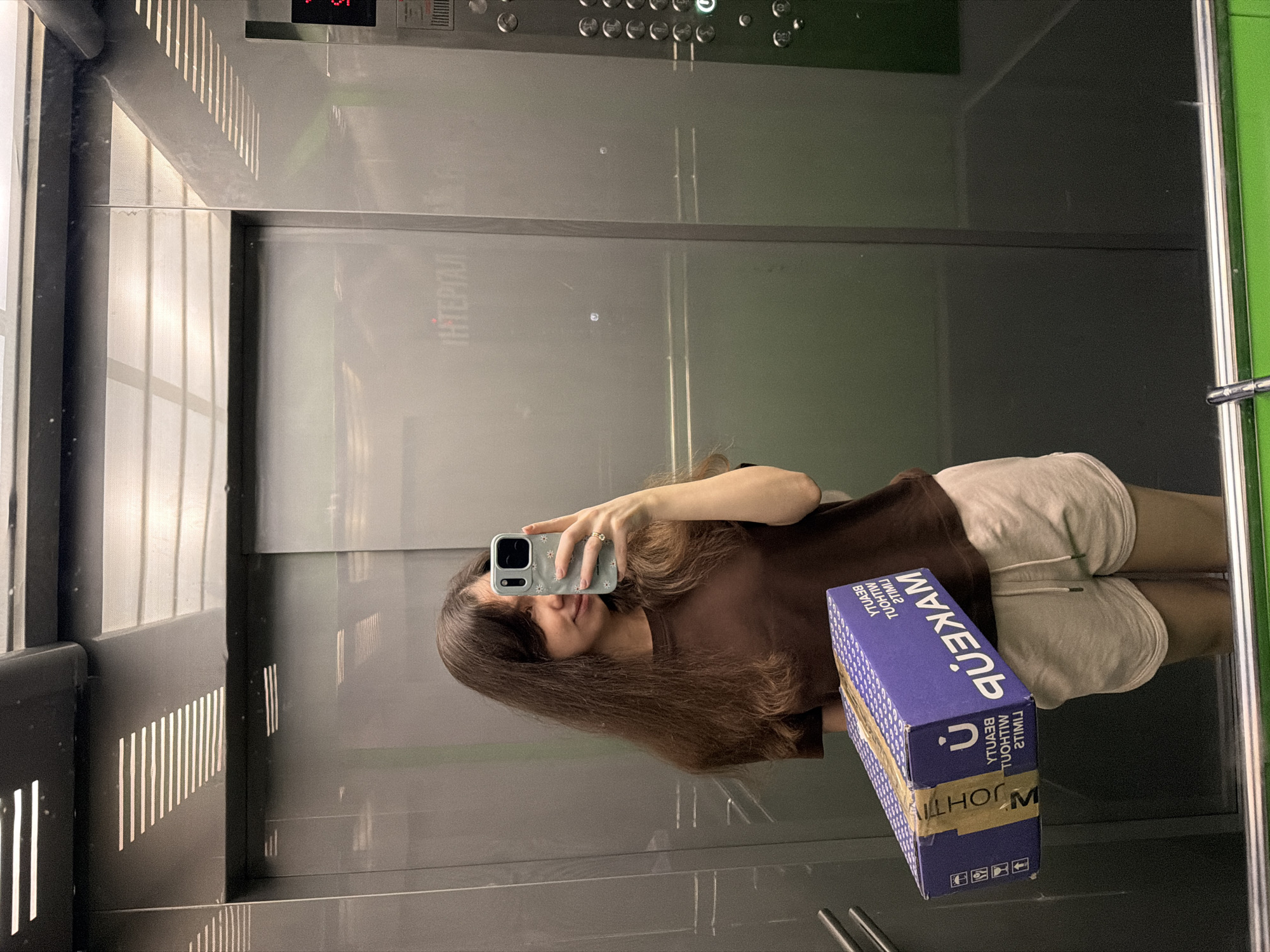 A young woman with long, dark brown hair stands in an elevator, taking a selfie with her phone. She is wearing a brown t-shirt and light-colored shorts. In her left arm, she holds a purple cardboard box with white lettering that reads "MAKEUP BEAUTY WITHOUT LIMITS". The elevator walls are reflective, showing the interior of the elevator car and the woman's reflection. The elevator control panel is visible on the right side of the image, with several buttons and a digital display. The woman appears to be in Kyiv, Ukraine, given the presence of Cyrillic text on the elevator wall.