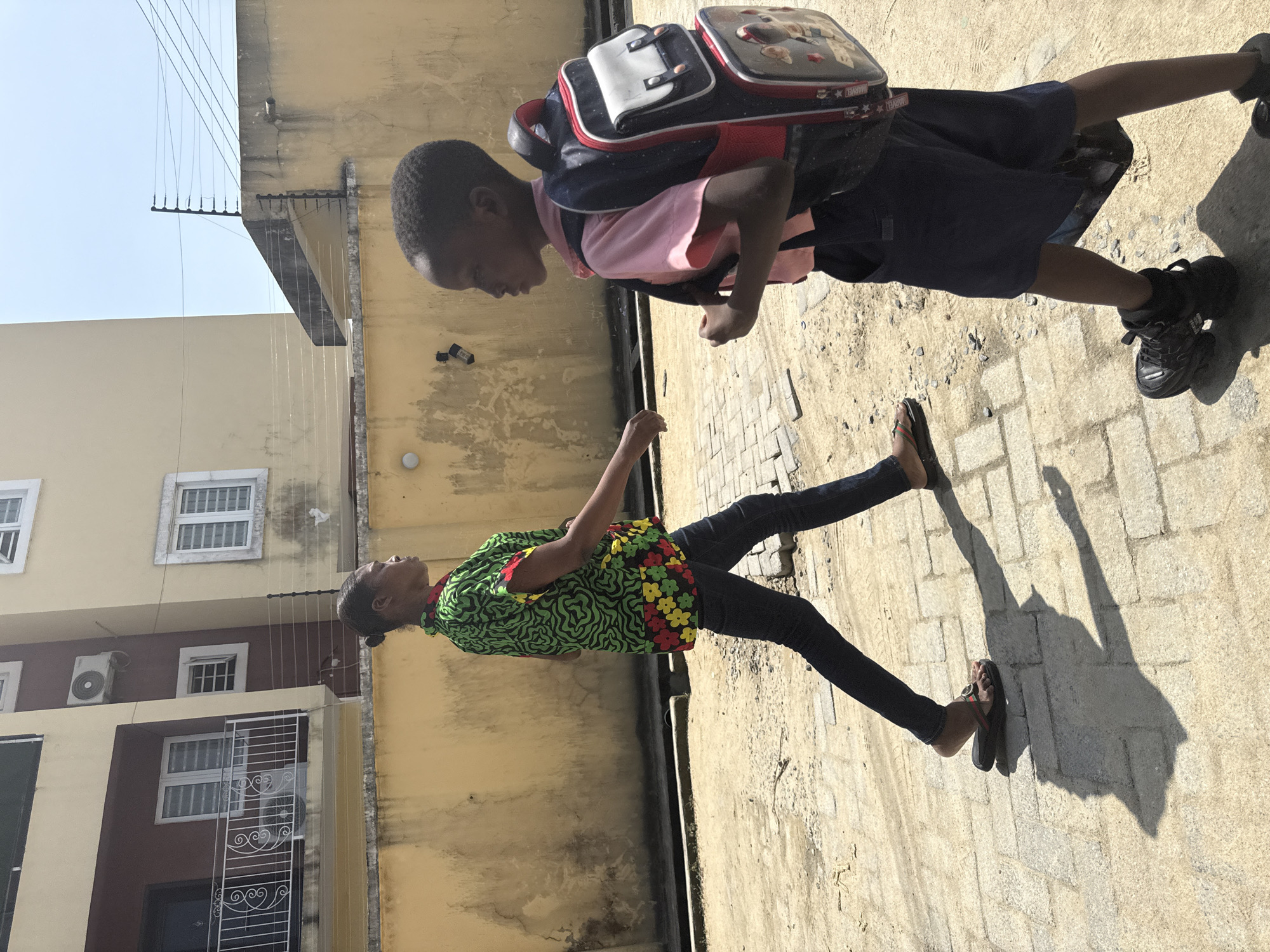In Ado, Nigeria, on a sunny day, a woman walks in profile from left to right, wearing a vibrant green shirt with a black abstract pattern and multi-colored floral accents at the hem, paired with dark jeans and flip-flops. She has her hair pulled back in a bun. To her right, a young boy walks towards the viewer, also in profile, wearing a pink collared shirt, dark shorts, black sneakers, and a large blue, white, and red backpack adorned with cartoon characters. They are walking on a paved surface with sandy patches, in front of a yellow, textured wall of a building. The background features a multi-story building with windows and balconies, and an electric fence runs along the top of the wall. The lighting suggests midday.