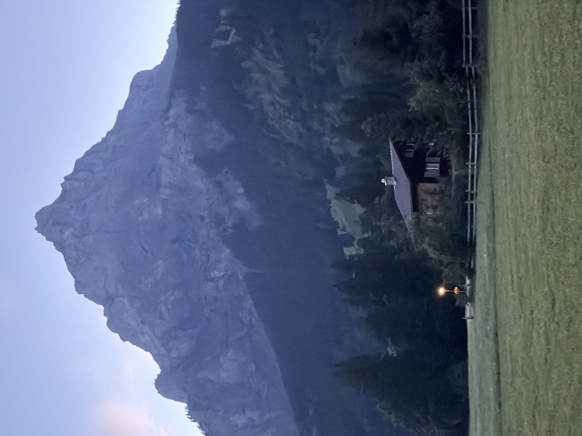 This is a photograph of a mountain range and a house in Kandersteg, Switzerland. The mountain is large and rocky, with snow on its peak. The house is a traditional Swiss chalet, with a wooden exterior and a steep, gabled roof. It is surrounded by trees and a wooden fence. The sky is a pale blue, and the overall mood of the photograph is peaceful and serene. The image appears to have been taken at dusk or dawn, as the light is soft and diffused. The house is illuminated by a single streetlamp.