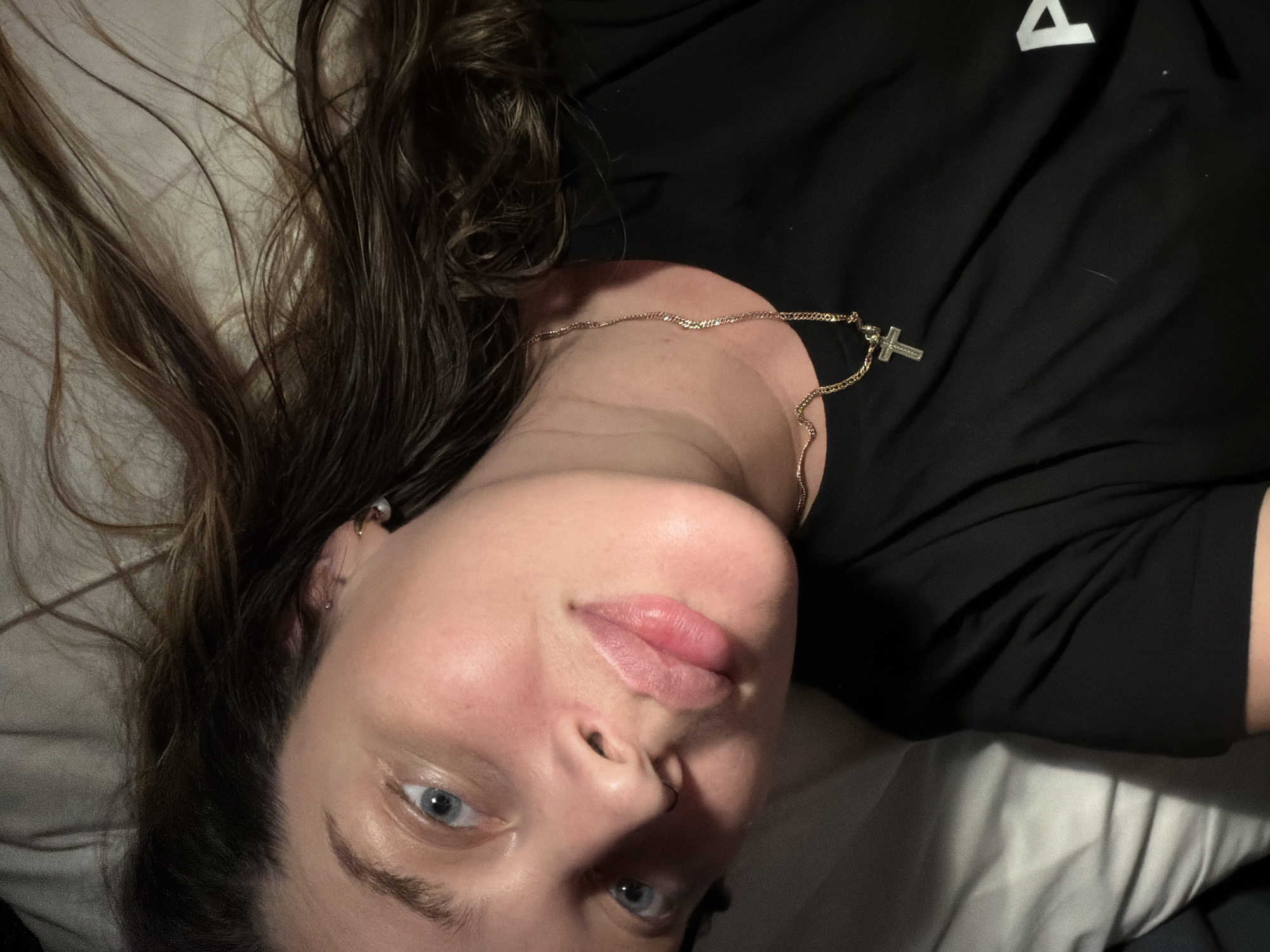 A close-up, rotated portrait shows a young adult with long, dark brown hair lying down, looking directly into the camera. Her hair is spread out on a light-colored pillow, suggesting an indoor setting, likely a bedroom, in Moscow, Russia. She has light skin, blue eyes, and full, light pink lips that are slightly parted. A small nose stud is visible on her right nostril, and multiple small earrings adorn her left ear. She is wearing a black t-shirt, which features a partial white logo resembling the top of an 'A' or a triangle on the shoulder. Around her neck is a gold chain necklace with a small silver cross pendant. The lighting is soft and subdued, indicating low light conditions, possibly evening or night. Her expression is neutral and calm.