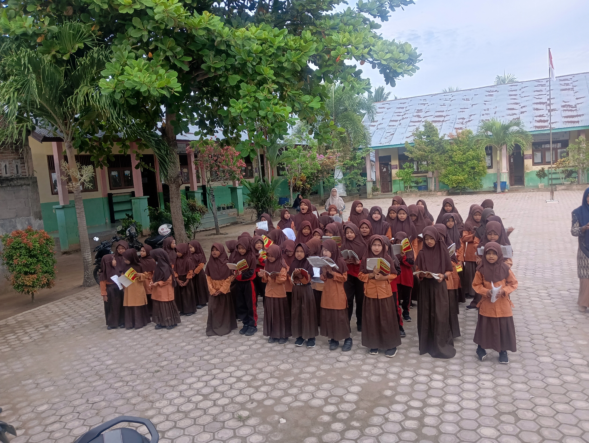 A group of young female students wearing brown hijabs and uniforms are gathered in an outdoor courtyard in front of a school building. Some students are holding books and appear to be reading or singing. The school building is painted yellow and green, with a corrugated metal roof. There are trees and a flagpole with an Indonesian flag visible. The ground is paved with hexagonal tiles. The scene appears to be in the city of Baratpayaitek, Indonesia.