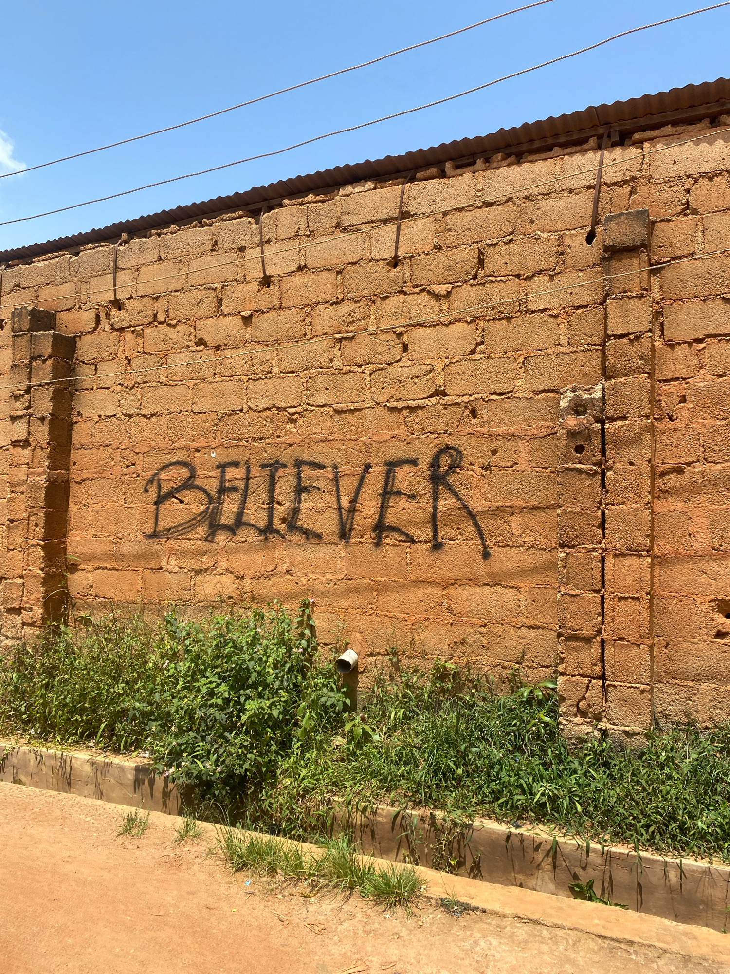 A high-angle, medium shot captures a brick wall adorned with black graffiti that reads "BELIEVER". The wall is constructed of large, rough, tan-colored bricks and is topped with corrugated metal sheeting. Below the wall, overgrown green vegetation lines a concrete drainage ditch that runs alongside a reddish-brown dirt road. A white pipe protrudes from the wall, disappearing into the foliage. The scene appears to be set in Jos, Nigeria, indicated by the sun-drenched environment and architectural style. The sky is a clear, pale blue.