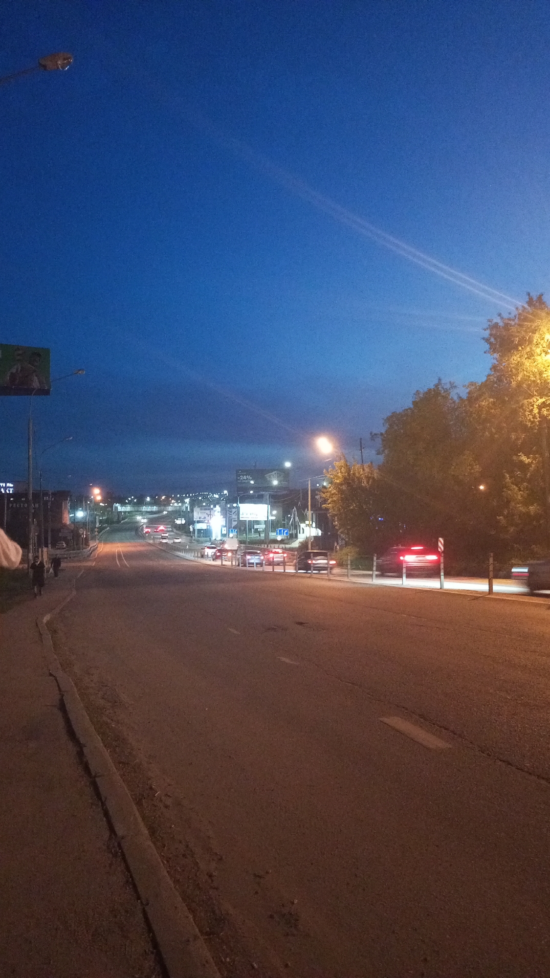 The image depicts an asphalt road in Юдино, Russia, at dusk or night. Street lights illuminate the road and surrounding trees on the right side of the frame. In the distance, the road inclines upwards, with multiple vehicles visible, indicated by headlights and taillights. A billboard stands to the left of the road. A pedestrian walks along the left sidewalk. A line of cars is stopped on the right side.
