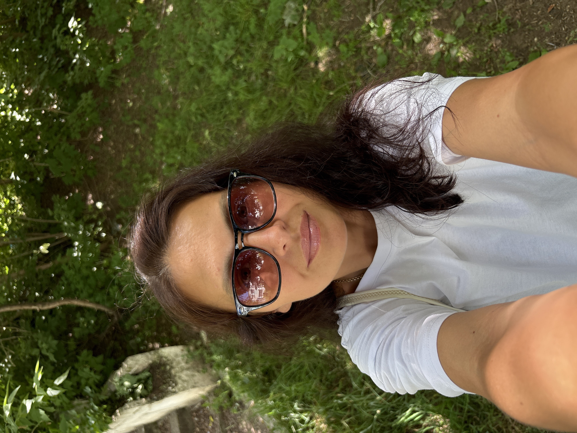 The media file is a selfie taken outdoors in Chaikovsky, Russia. A woman with long, dark brown hair is the primary subject. She is wearing a white t-shirt and sunglasses with dark, reflective lenses that show a blurred image of trees. The background consists of lush green grass and foliage, suggesting a natural or park-like setting. The lighting indicates it is daytime, likely sunny, casting shadows and highlighting the textures of the leaves and grass. The woman's face is tilted upwards, and she is looking towards the camera. Her lips are slightly parted, and she appears relaxed and content. A faint outline of a gold chain necklace is visible around her neck. The overall scene evokes a sense of casual enjoyment of an outdoor environment. There is no visible text in the media file.