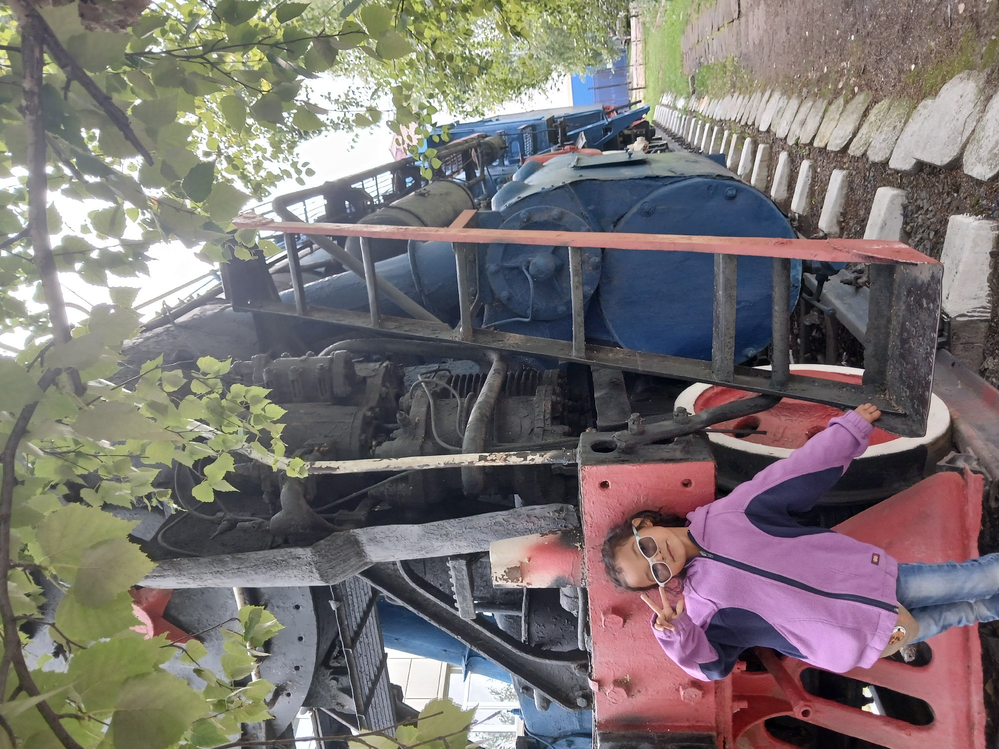 A young girl is posing next to a train engine. The girl is wearing sunglasses and a purple and blue jacket. She is standing with her left arm raised and making a peace sign with her fingers. The train engine is red and blue and has a ladder attached to it. The train is on tracks, and there are trees and foliage in the background. The scene is likely located in Щучинск қаласы әкімдігі, Kazakhstan.