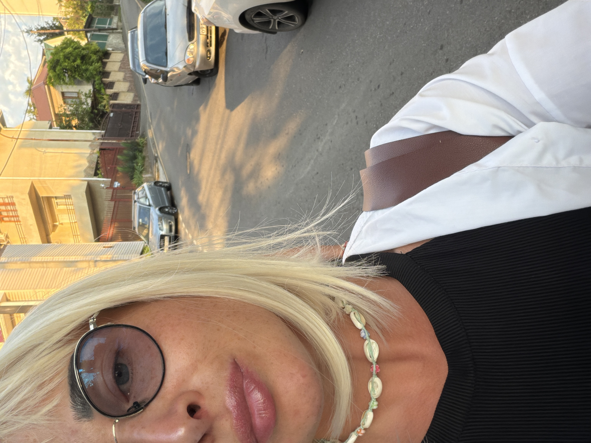 A medium shot captures a woman with blonde hair and sunglasses as she takes a selfie on a street in Sochi, Russia. The street slopes upwards and is lined with buildings on the left and trees on the right. Several cars are parked along the street, and a few are in motion. The woman is wearing a black top, a white unbuttoned shirt, and a necklace made of shells and small colorful beads. Her lips are slightly parted and appear to be glossed. The lighting suggests it is late afternoon, with long shadows cast across the road.