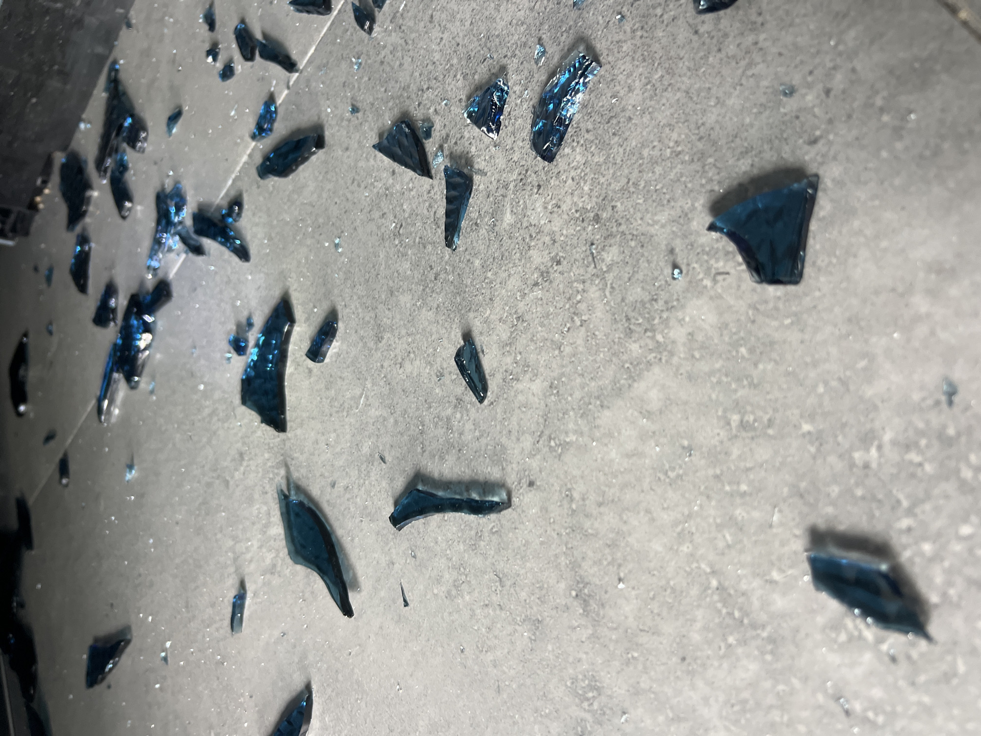 The image depicts numerous shards of blue glass scattered across a textured, light gray surface that appears to be tile or concrete. The glass fragments vary in size and shape, with some being thin and sharp, while others are thicker and more irregular. The surface also contains a scattering of smaller, glittering particles, possibly dust or tiny fragments of glass. In the background, a dark, out-of-focus object is visible, suggesting an indoor environment. There is no indication of the specific location within Sochi, Russia, beyond it being an indoor area with a tiled or concrete floor. No people, actions, or interactions are visible in the image.