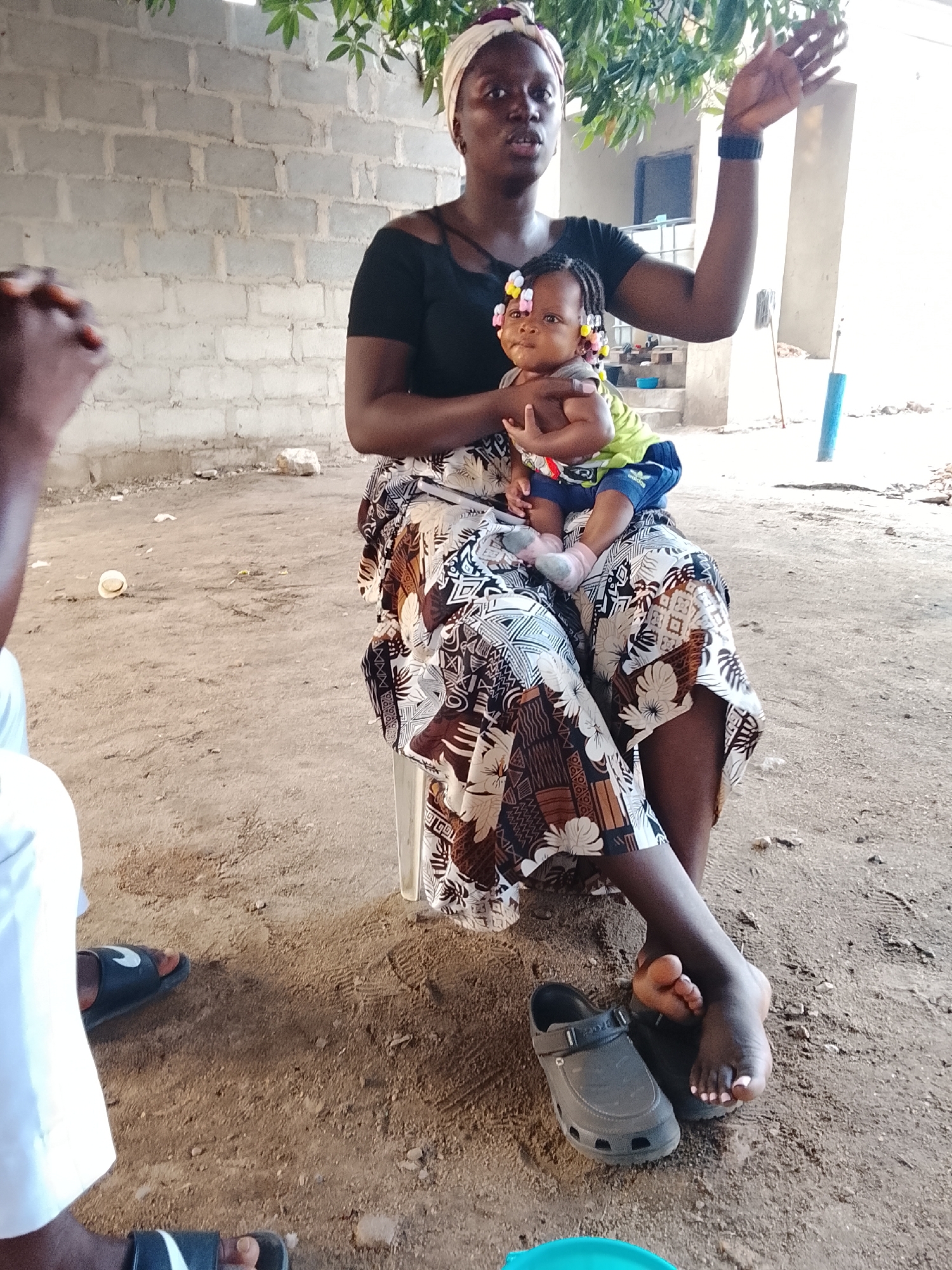 The image shows a woman seated outdoors with a baby on her lap. The woman is wearing a headwrap, a black top, and a patterned skirt. The baby has colorful hair beads and is wearing a lime green top with blue shorts. The woman is raising her left arm in a gesture of speaking or gesturing. The setting appears to be a yard with a dirt ground and a partially visible cinder block wall in the background. One person is partially shown in the bottom left of the image. A pair of gray crocs are present to the left of the woman's right foot. The location may be Akwanga, Nigeria.