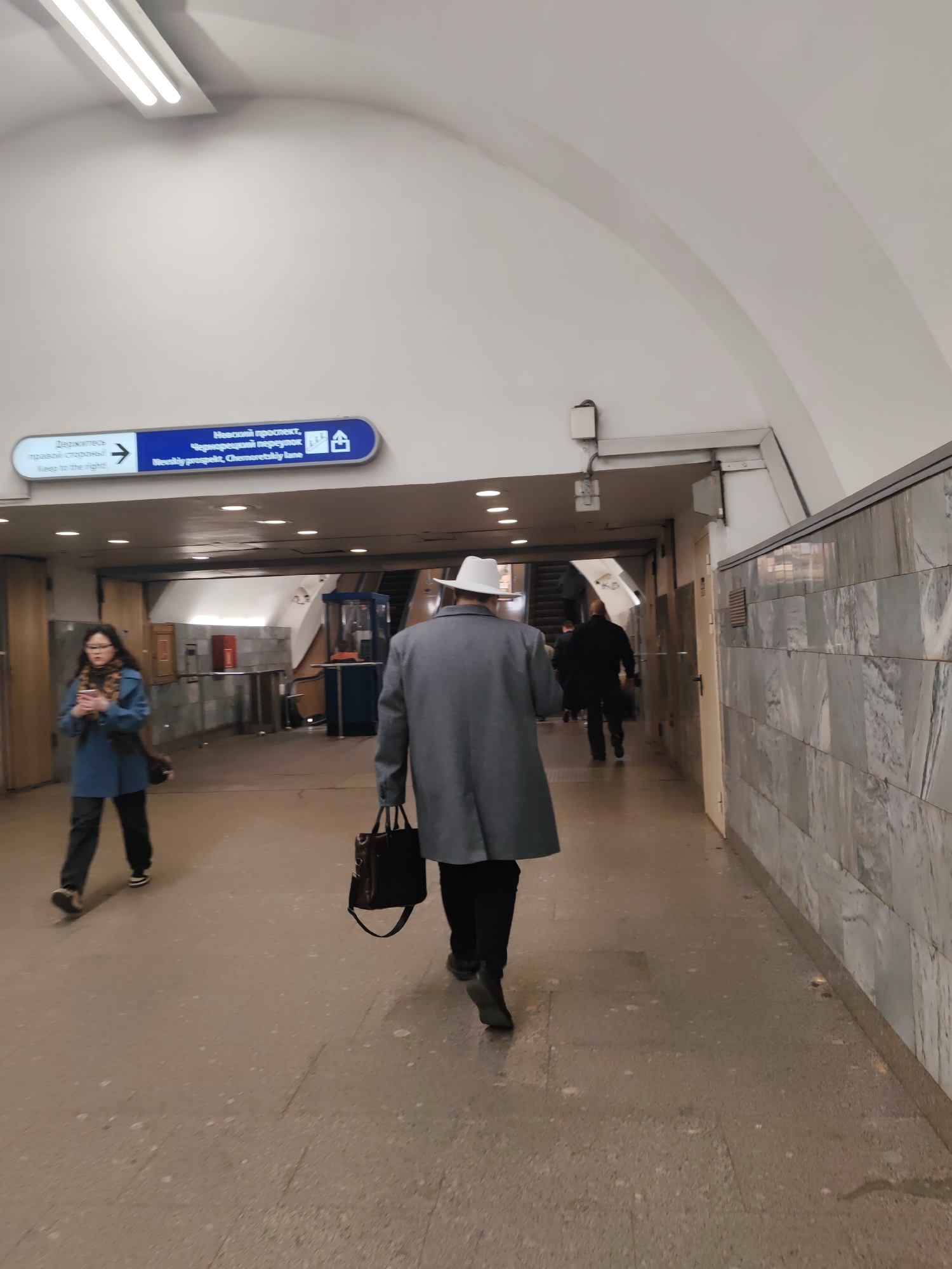 This vertical shot captures an underground pedestrian passage, likely within a metro station in Saint Petersburg, Russia. The scene depicts several commuters moving through the brightly lit, arch-ceilinged walkway.
In the foreground, a person seen from behind is walking away from the camera. They wear a light grey overcoat, dark trousers, and a distinctive white fedora-style hat, carrying a dark leather bag in their right hand. To the left, another individual, a woman with long dark hair and glasses, is walking towards the camera. She wears a blue coat over a patterned scarf and appears to be looking at her smartphone. Further down the passage, other figures are visible, mostly as silhouettes, heading in the same direction as the person in the white hat, towards what appears to be an escalator or a set of turnstiles.
The passage features a light-colored, possibly concrete or terrazzo, floor and a uniformly white, arched ceiling equipped with long fluorescent light fixtures. The right wall is clad in grey and white marble or stone tiles, while the left wall is plainer, holding a prominent blue and white directional sign.
The visible text on the sign, in both Russian and English, reads:
"Держитесь правой стороны! Keep to the right!"
"Невский проспект, Чернорецкий переулок Nevskiy prospekt, Chernoretskiy lane"
Accompanying icons on the sign indicate stairs/escalator and possibly a building or exit.
The overall atmosphere suggests a typical commuting moment in an urban underground environment. The artificial lighting indicates an indoor setting where the time of day is not a factor.