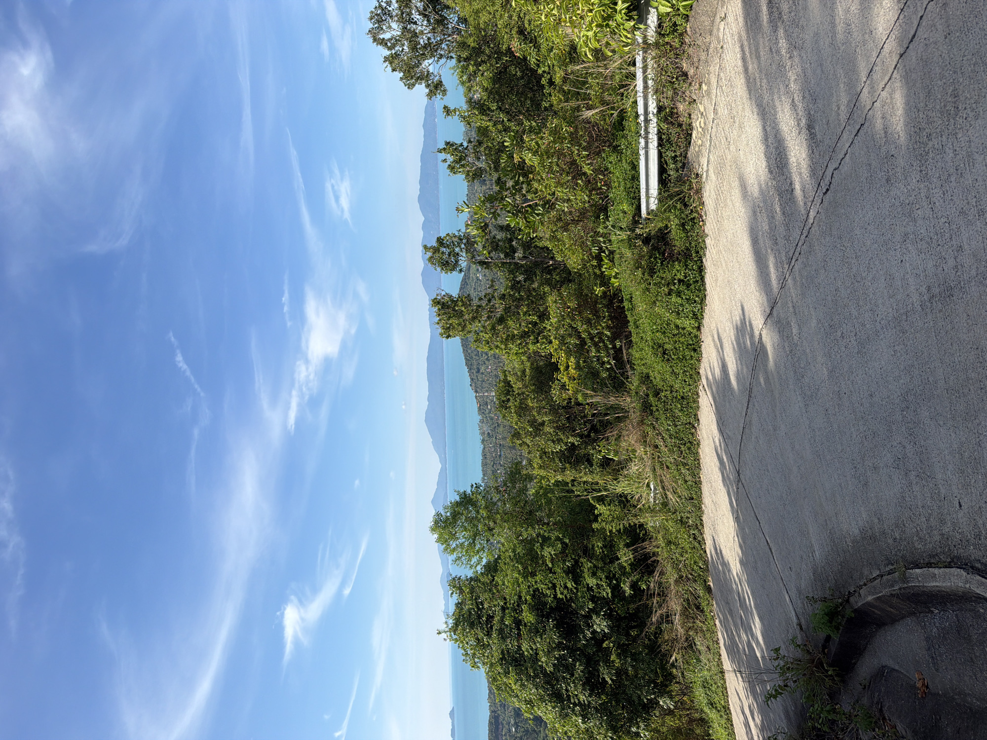 A scenic view of the ocean and mountains is captured from a paved road that curves to the left. The road is in the foreground, with shadows from trees cast across its surface. To the right of the road, a white metal guardrail is visible, separating the road from a dense green foliage. In the background, lush green trees cover a hillside, with the turquoise ocean stretching out beyond them. A mountain range can be seen in the distance, under a bright blue sky with scattered white clouds. The location is identified as Baan Tai, Thailand. There are no people or animals visible in the image.