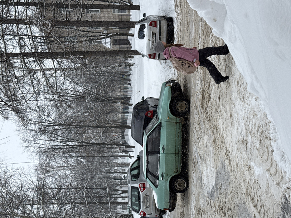 This image depicts a snowy urban scene in Chaikovsky, Russia, characterized by overcast weather and visible snowfall. In the lower right, a young girl, wearing a grey beanie, a pink winter jacket, dark pants, and dark boots, walks from right to left, carrying a light tan backpack and a pink object in her left hand. The ground where she walks is a mix of compacted snow and slush.
Behind the girl and extending to the left, a slushy roadway is present. Four vehicles are visible. In the foreground on the roadway, a light green sedan (likely a Lada Samara/Sputnik series, possibly a VAZ-21099) with a damaged front bumper is parked or stopped, facing left. Behind it, a silver SUV with Russian license plate "T274PK 159" is parked. To the right of the green sedan, a black hatchback or sedan is partially visible, facing right. Further right and behind these, a white Skoda Octavia sedan is parked in a snowdrift, facing right, with its rear window covered in snow and license plate "O584KM 159" partially visible.
The background features numerous bare deciduous trees covered in snow, indicating winter. A dark metal fence runs horizontally through the mid-ground. Beyond the fence on the right, a multi-story building constructed of light-colored bricks is partially visible through the trees. The entire scene is covered in snow, with deeper drifts along the edges of the cleared areas.
