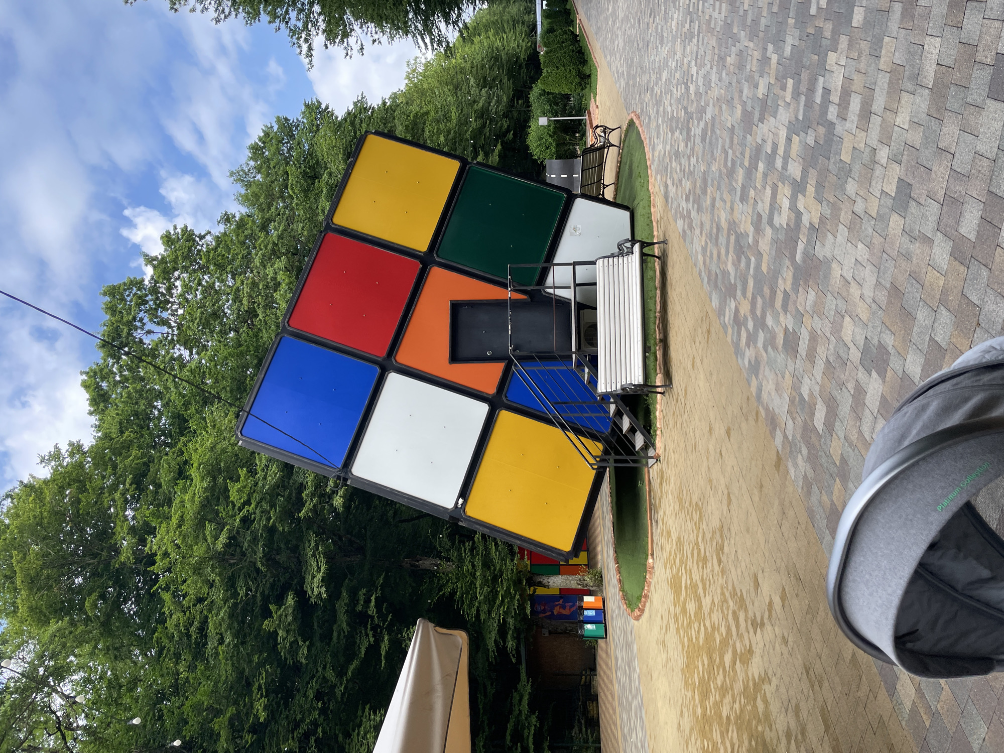The image captures a large, tilted Rubik's Cube structure situated in an outdoor park-like setting. The cube is positioned at an angle and features a black door and stairs leading to it. White benches are placed in front of the structure, and a paved walkway with a patterned brick design extends into the background. Lush green trees form a dense backdrop, and the sky above is partly cloudy. In the foreground, a portion of a gray baby stroller is visible. The scene appears to be a public space, possibly a recreational area or a theme park attraction. It is noted that the location is the city of Stavropol, Russia.