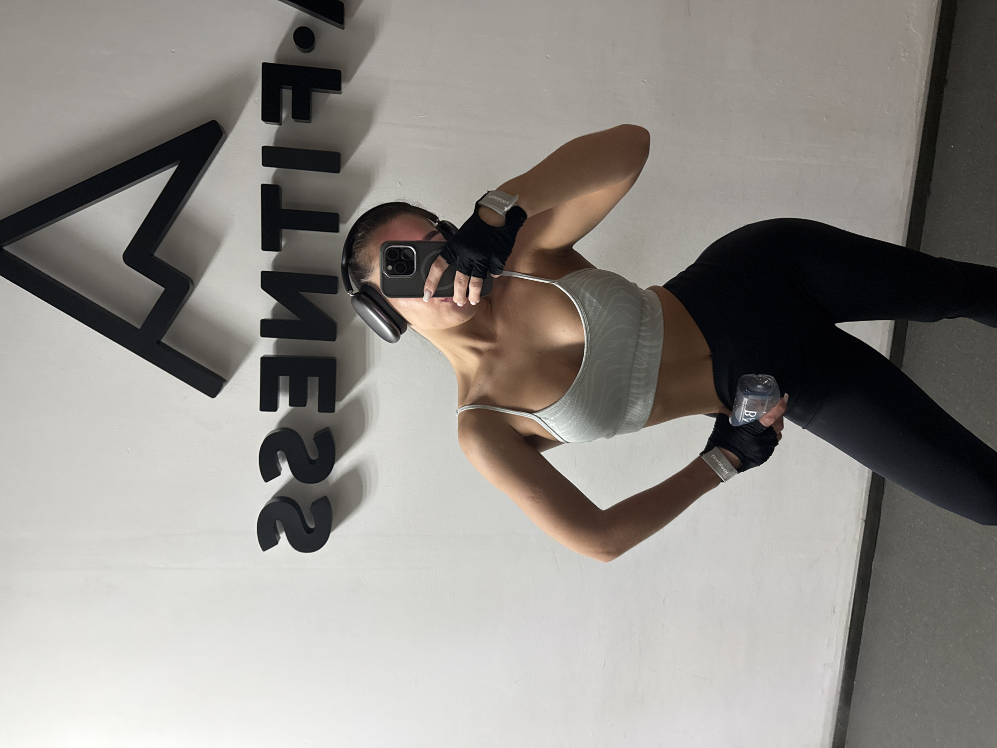 A woman is taking a selfie indoors. She is wearing a light-colored sports bra, black leggings, black gloves with wristbands, and headphones. She is holding a phone in one hand and a water bottle in the other. Behind her is a white wall with black lettering that appears to be a logo or name of a gym. A partial view of a black textured floor is also visible in the right edge of the image. The gym could be located in the city of Kazan, Russia.