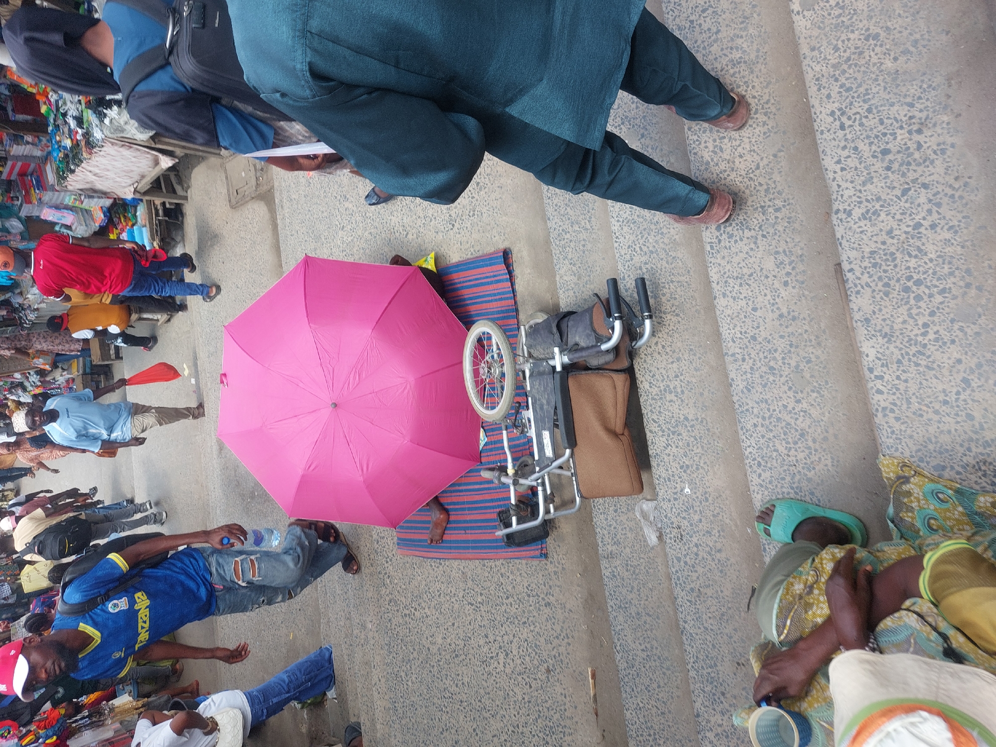 The scene is an outdoor street view in Dar es-Salaam, Tanzania. A pink umbrella shades a folded wheelchair resting on a striped mat. Nearby, an individual sits on the steps, covered in patterned fabric, with a light-colored hat and a blue cup. Pedestrians, including one wearing a blue Tanzania-branded jersey and another in a dark green outfit, are visible passing by, adjacent to market stalls with assorted goods.