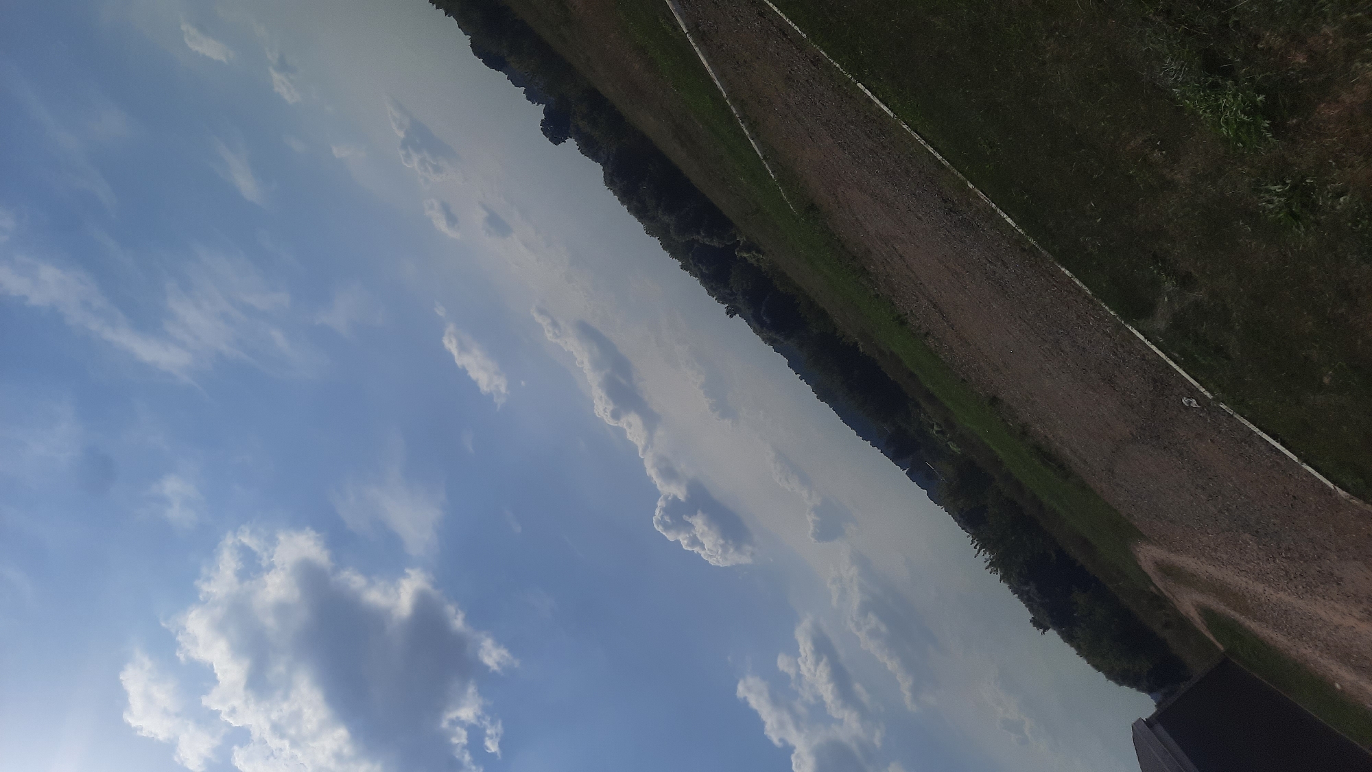 This is an image of a rural landscape in Shklov, Belarus. The photo is taken at an angle, looking upwards towards a sky with scattered clouds and a blue sky. Below the sky is a dark line of trees, which appears to be a forest. In front of the trees, there is a field that transitions from dirt to grass. A white line runs diagonally across the field. On the left side of the image, there is a portion of a dark-colored building visible. The overall scene suggests an outdoor environment, possibly a park or a field on the outskirts of town.