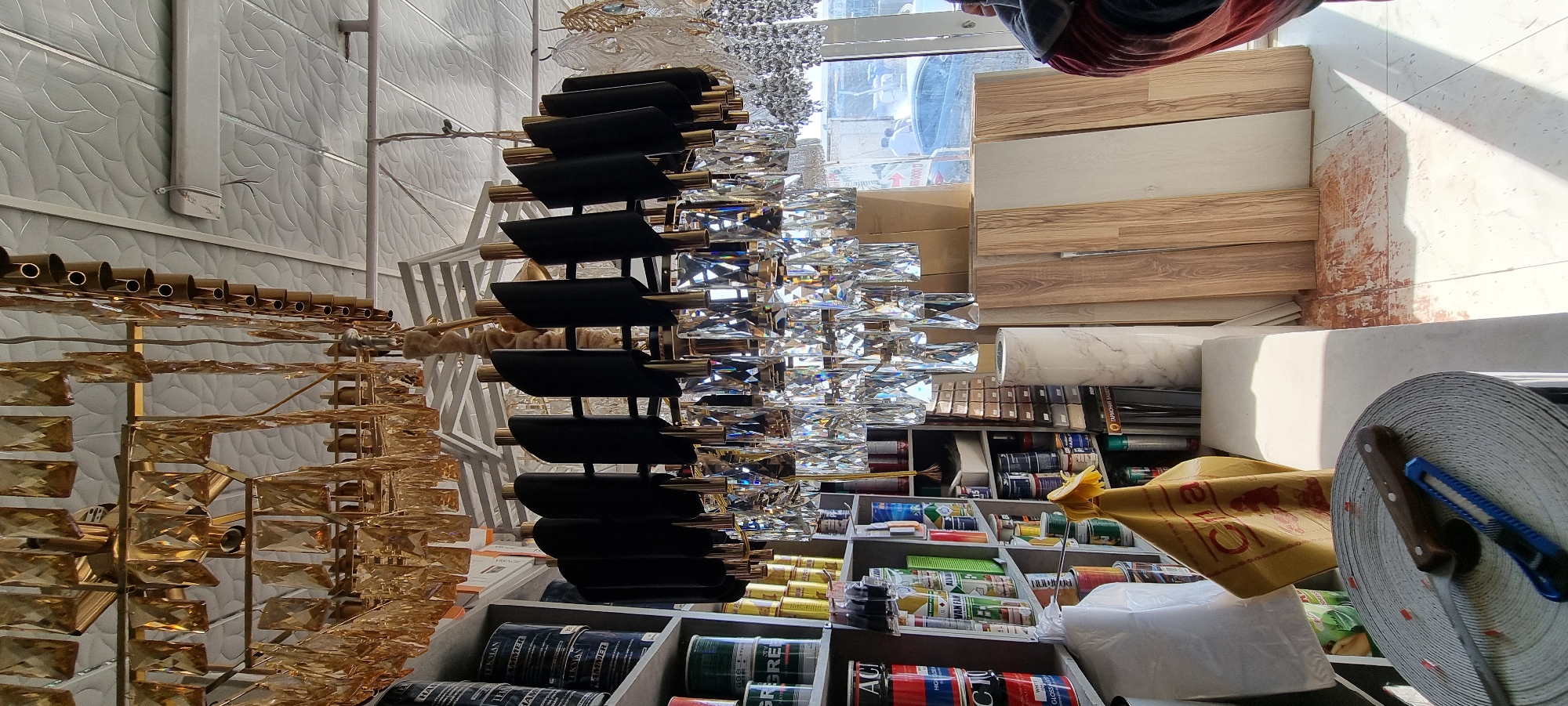 The image depicts the interior of a retail establishment, likely a store selling home improvement or decorative items, located in the city of Bokhtar, Tajikistan. The primary subject is a large, ornate chandelier hanging from the ceiling. This chandelier features multiple tiers with amber-colored rectangular crystals and black, curved decorative elements. Below the chandelier, on shelves, are various products, including cans of paint or similar containers, rolls of material, and other miscellaneous items. To the right of the shelves, there are stacks of what appear to be wood planks or laminate flooring, and a marble-patterned roll of material. In the foreground, a roll of tape or similar material is visible, with a utility knife and a standard knife resting on top. The environment suggests a commercial setting with fluorescent lighting from the ceiling and natural light entering from a window or doorway in the background, through which a vehicle and two individuals can be partially seen. The overall impression is one of a busy retail space displaying a variety of goods.