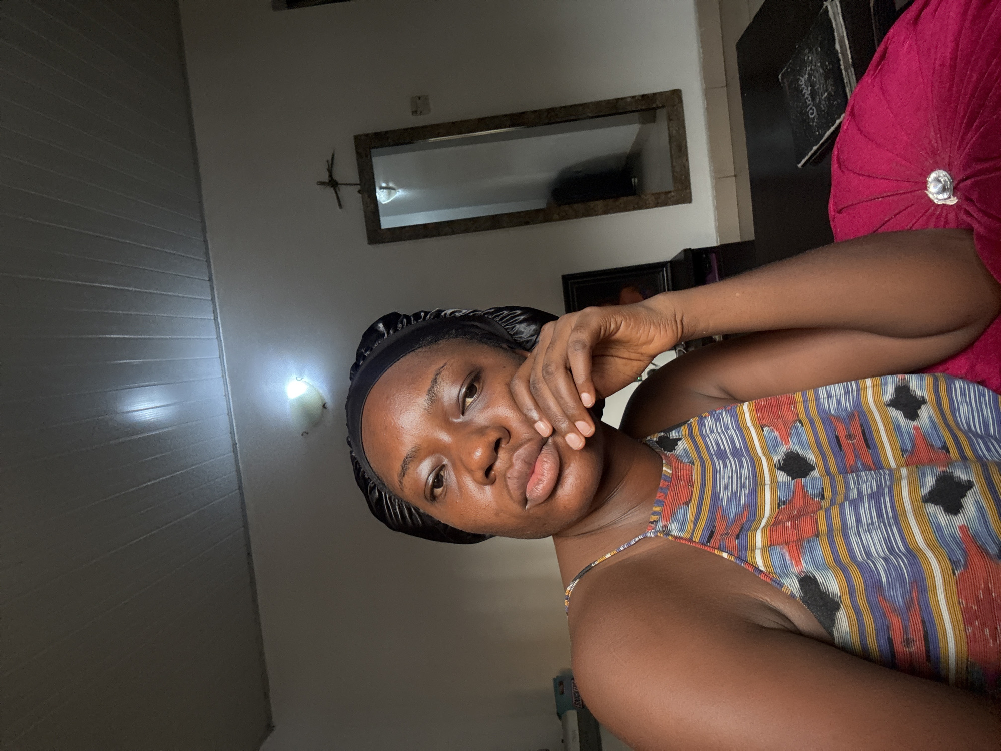 A young dark-skinned woman poses for a close-up photo, likely a selfie, while reclining indoors. She is looking directly at the camera with a neutral, slightly pensive expression, her lips slightly parted. Her right hand rests gently against her chin and cheek. She wears a black satin bonnet and a vibrant, patterned sleeveless top with thin straps, featuring vertical stripes and geometric shapes in blue, red, orange, black, and white – suggestive of an Ankara print.
The setting appears to be a domestic interior, possibly a bedroom or living room, consistent with a location in Abuja, Nigeria. Behind her, a light-colored wall features horizontal paneling on the left. Above her head, a rectangular mirror with a dark frame reflects indistinct shapes. To the right of the mirror, a dark, angular wall decoration is visible. On the right side of the frame, a plush, dark red or magenta pillow with a central button detail rests against a dark piece of furniture, which might be a headboard or sofa back. Some faint, illegible text is visible on the dark furniture next to the pillow. Artificial light sources are visible, casting a glow on parts of the wall and ceiling, suggesting the photo was taken in the evening or in an area with dim natural light. The overall mood is calm and relaxed.